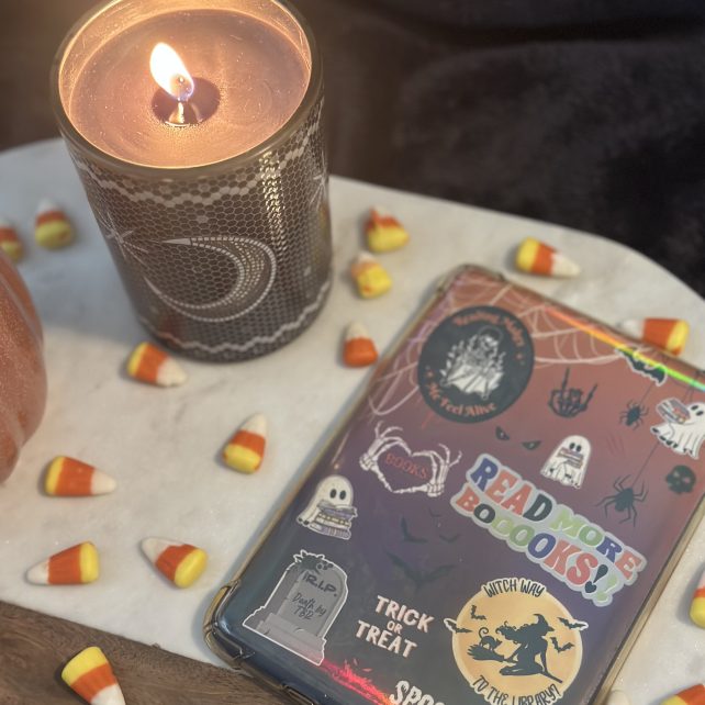 A lit black and white honeycomb-print candle next to a holographic Halloween Kindle insert featuring ghost, spider, tombstone, and witch stickers with "Read More Boooooks" and "Spooky Vibes" text, surrounded by candy corn on a marble and wood tray.