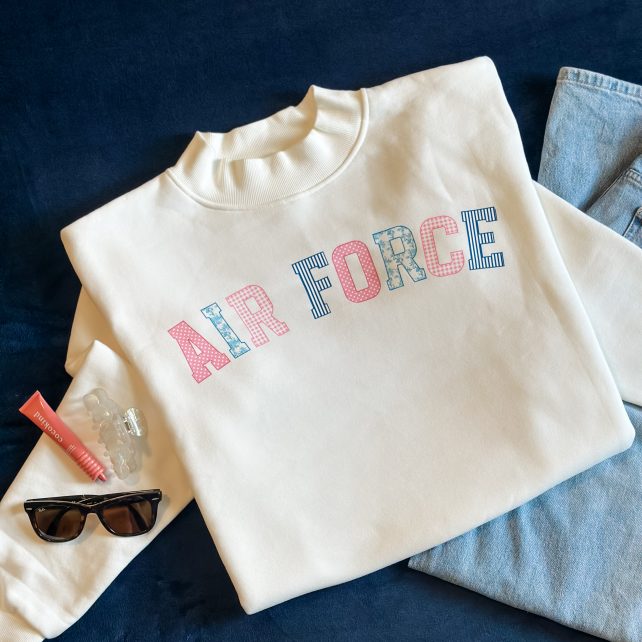 White mockneck sweatshirt with "Air Force" in pink and blue grandmillennial patchwork lettering, styled flat with jeans, sunglasses, and a hair clip on a navy background