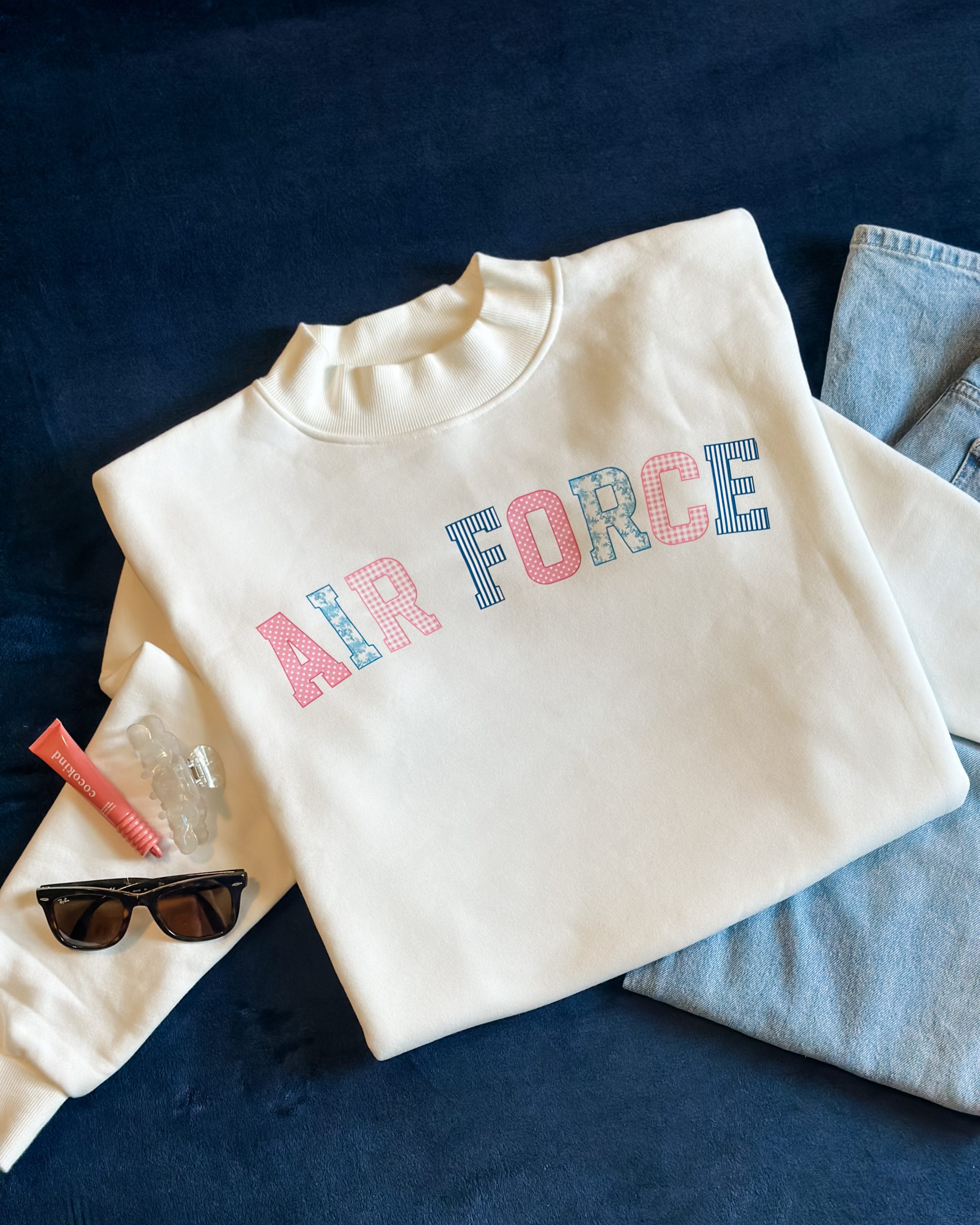 White mockneck sweatshirt with "Air Force" in pink and blue grandmillennial patchwork lettering, styled flat with jeans, sunglasses, and a hair clip on a navy background
