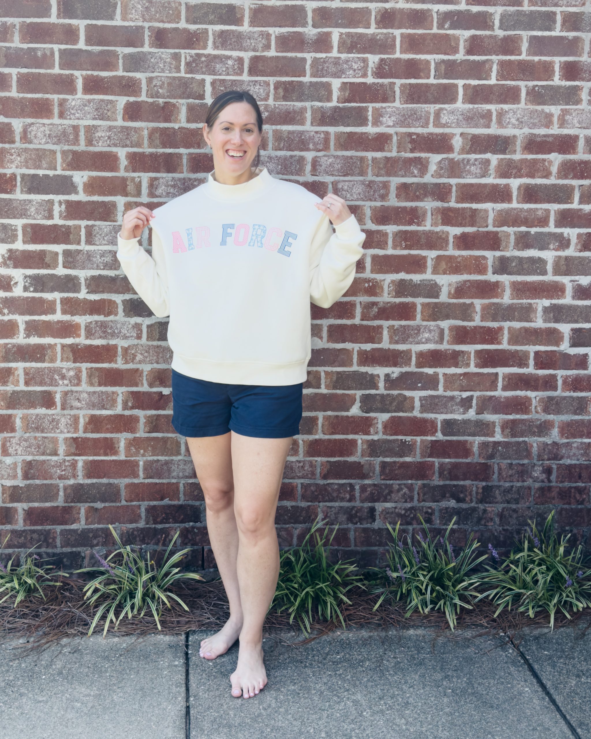 Woman smiling and holding out her white mockneck sweatshirt with "Air Force" in pink and blue grandmillennial patchwork lettering, standing in front of a brick wall
