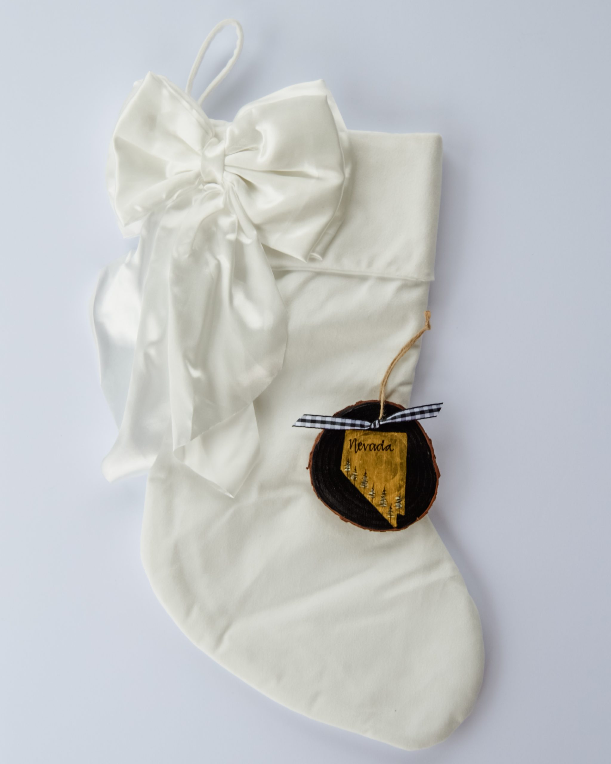 Hand-painted wood slice ornament of Nevada with a gold state silhouette, snow-covered trees, and the state name hand-lettered in black, tied with white buffalo plaid ribbon on a white velvet Christmas stocking