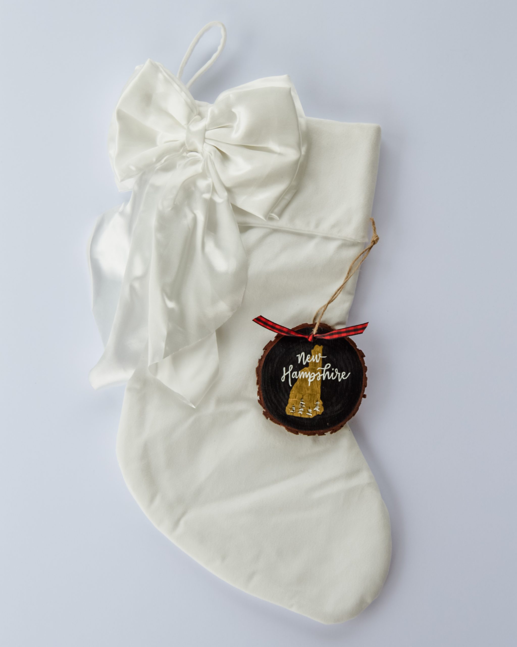 Hand-painted wood slice ornament of New Hampshire with a gold state silhouette, snow-covered trees, and the state name hand-lettered in white, tied with red buffalo plaid ribbon on a white velvet Christmas stocking