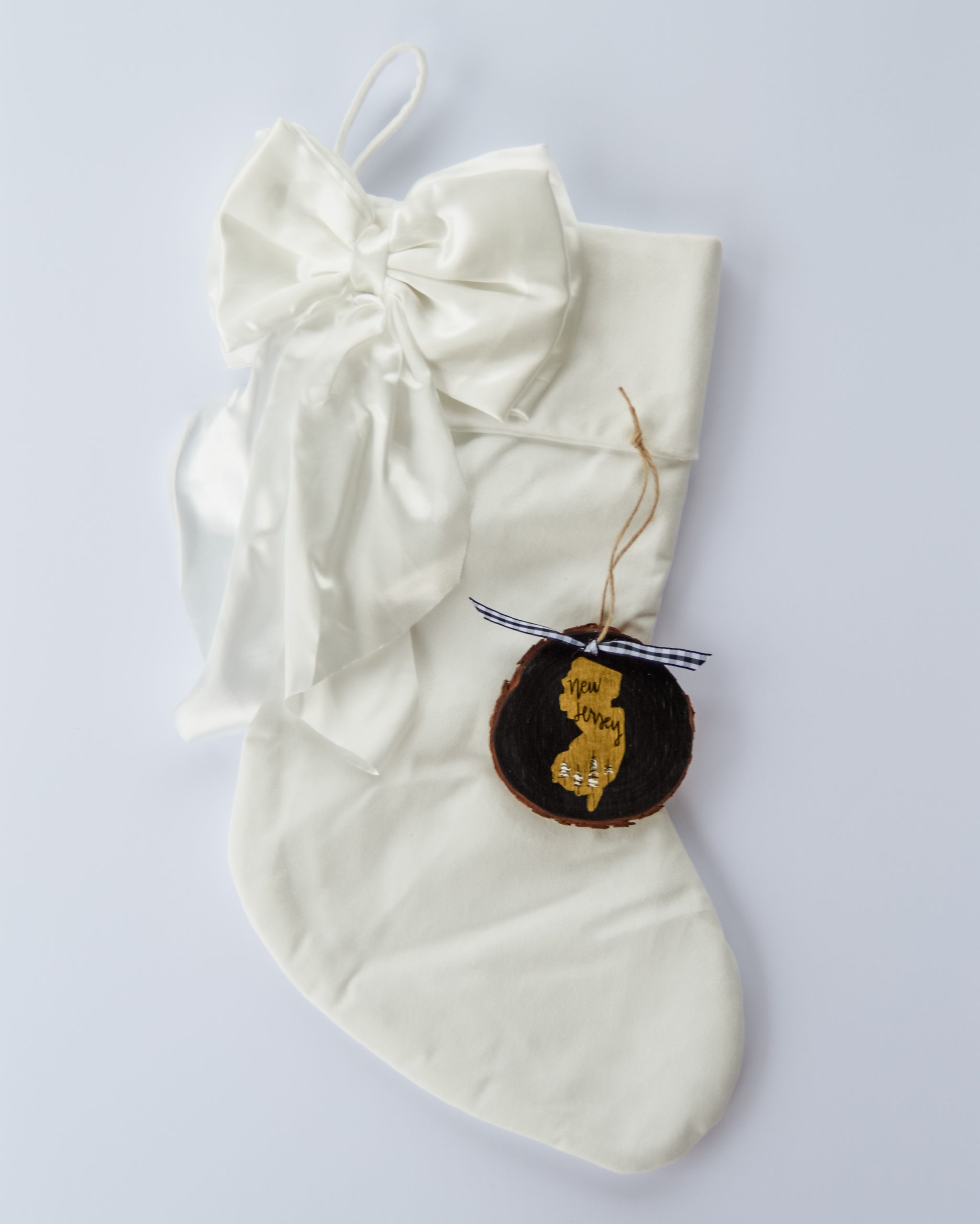 Hand-painted wood slice ornament of New Jersey with a gold state silhouette, snow-covered trees, and the state name hand-lettered in black, tied with white buffalo plaid ribbon on a white velvet Christmas stocking