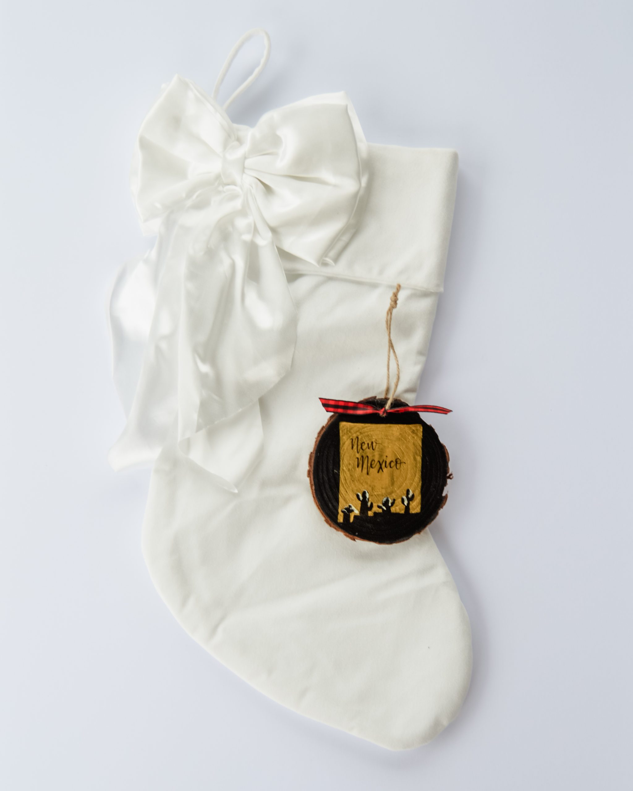 Hand-painted wood slice ornament of New Mexico with a gold state silhouette, snow-covered cacti, and the state name hand-lettered in black, tied with red buffalo plaid ribbon on a white velvet Christmas stocking
