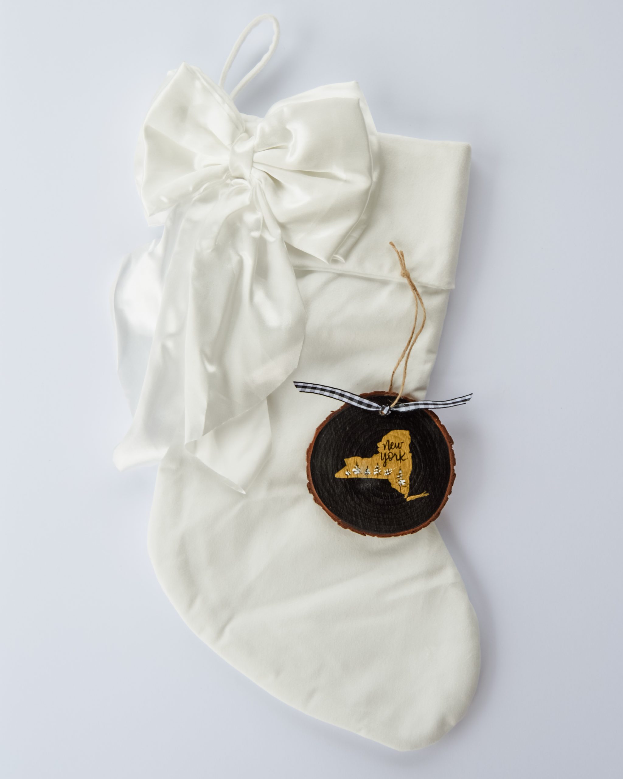 Hand-painted wood slice ornament of New York with a gold state silhouette, snow-covered trees, and the state name hand-lettered in black, tied with white buffalo plaid ribbon on a white velvet Christmas stocking