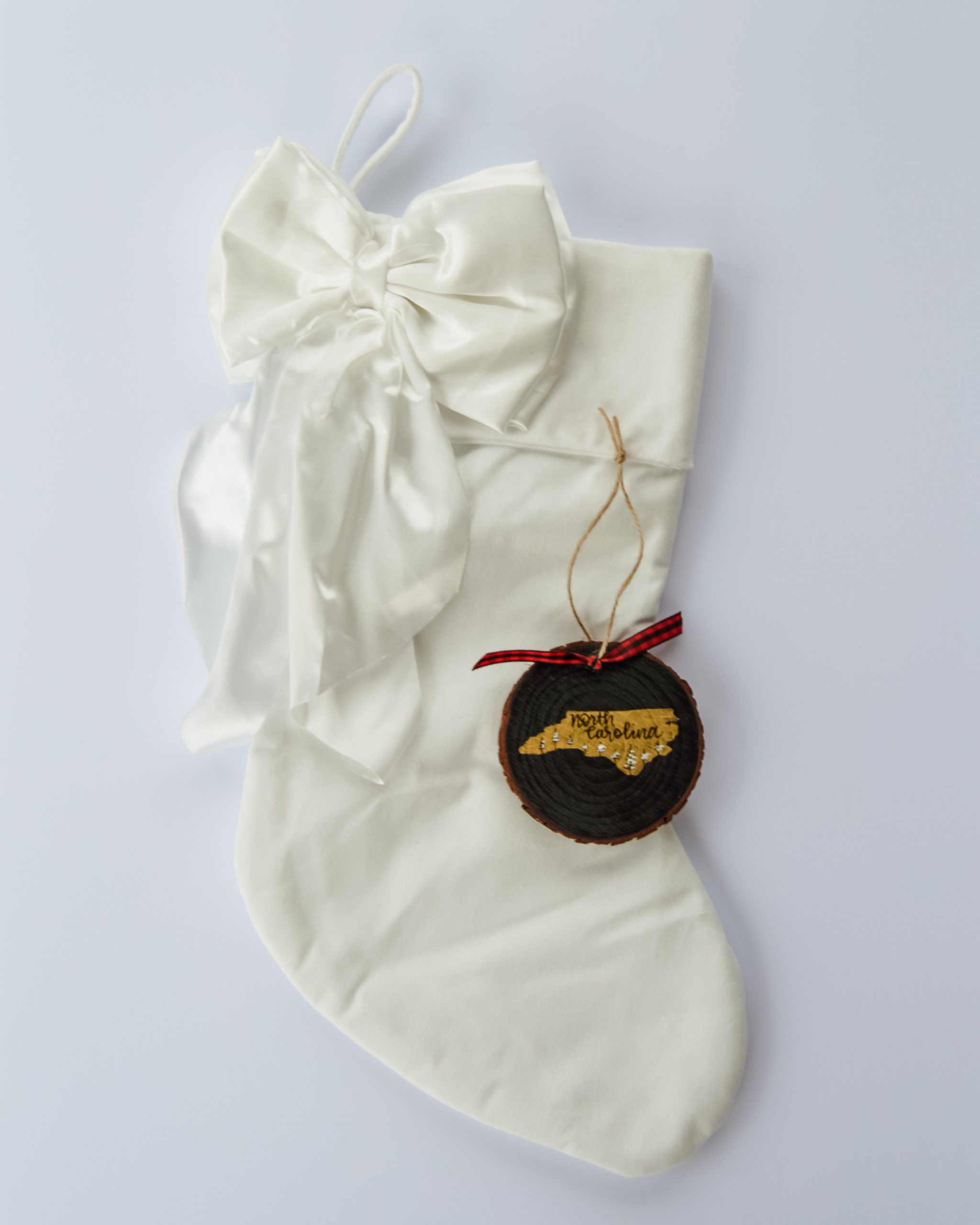 Hand-painted wood slice ornament of North Carolina with a gold state silhouette, snow-covered trees, and the state name hand-lettered in black, tied with red buffalo plaid ribbon on a white velvet Christmas stocking