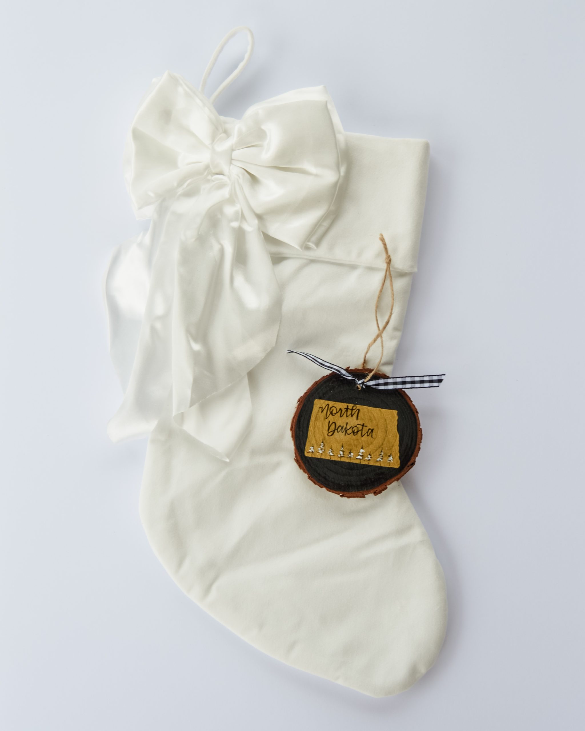Hand-painted wood slice ornament of North Dakota with a gold state silhouette, snow-covered trees, and the state name hand-lettered in black, tied with white buffalo plaid ribbon on a white velvet Christmas stocking
