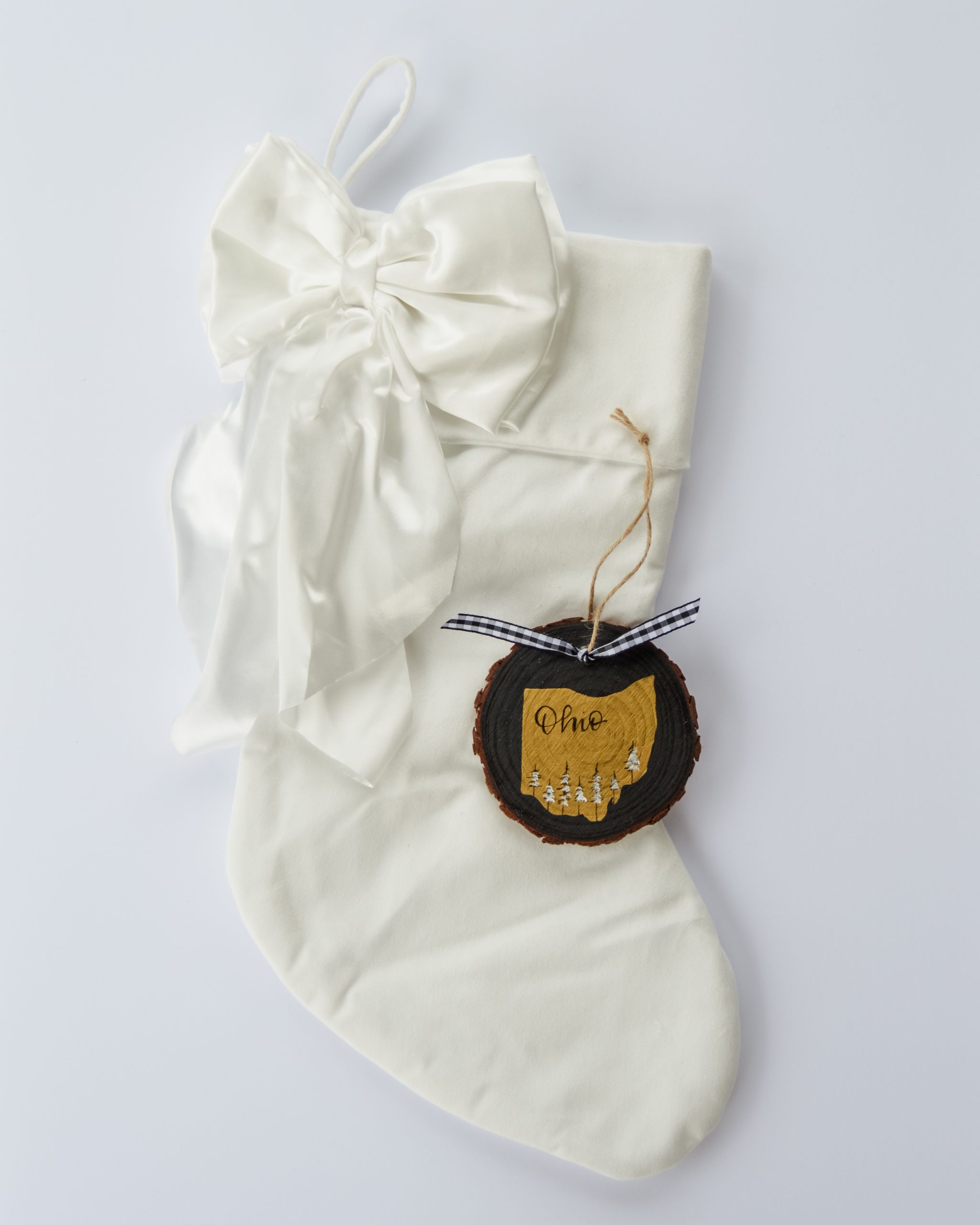 Hand-painted wood slice ornament of Ohio with a gold state silhouette, snow-covered trees, and the state name hand-lettered in black, tied with white buffalo plaid ribbon on a white velvet Christmas stocking