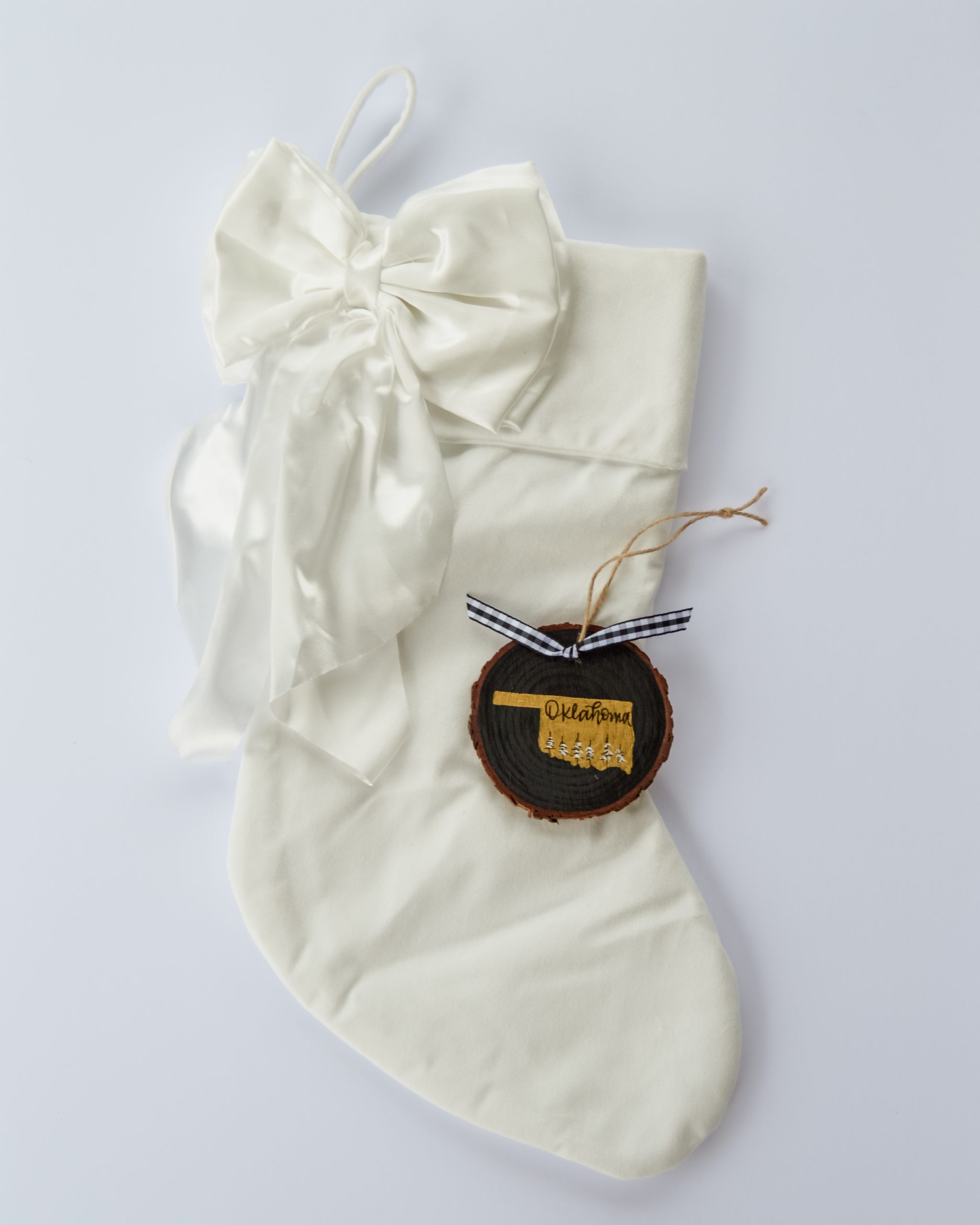 Hand-painted wood slice ornament of Oklahoma with a gold state silhouette, snow-covered trees, and the state name hand-lettered in black, tied with white buffalo plaid ribbon on a white velvet Christmas stocking