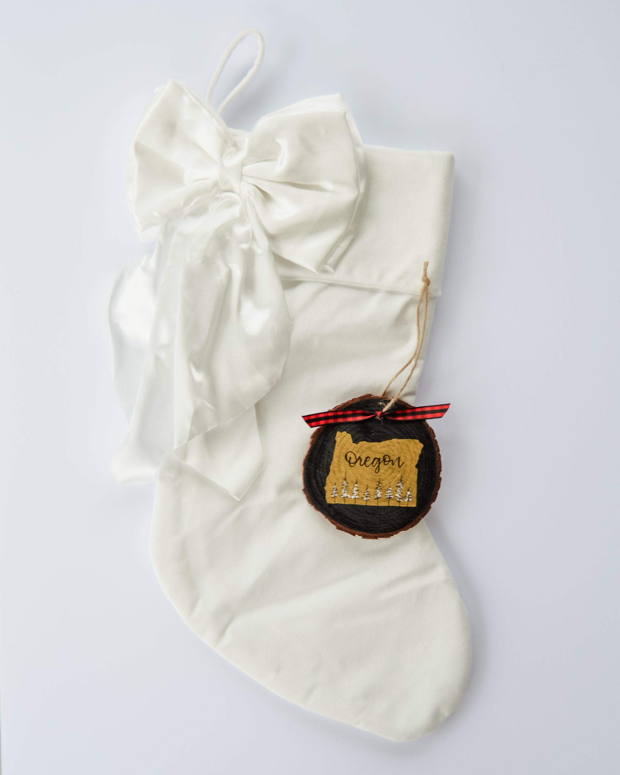 Hand-painted wood slice ornament of Oregon with a gold state silhouette, snow-covered trees, and the state name hand-lettered in black, tied with red buffalo plaid ribbon on a white velvet Christmas stocking