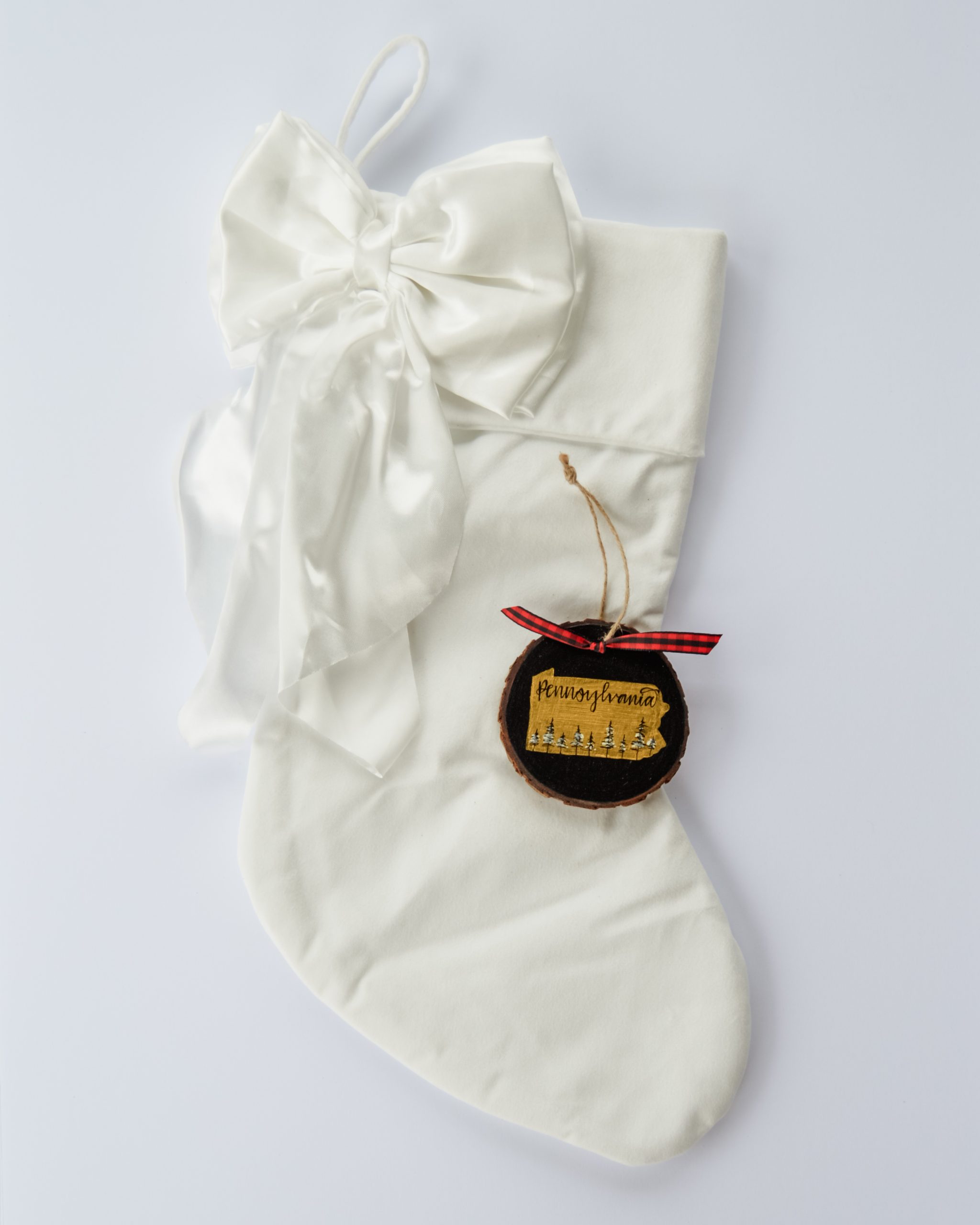 Hand-painted wood slice ornament of Pennsylvania with a gold state silhouette, snow-covered trees, and the state name hand-lettered in black, tied with red buffalo plaid ribbon on a white velvet Christmas stocking