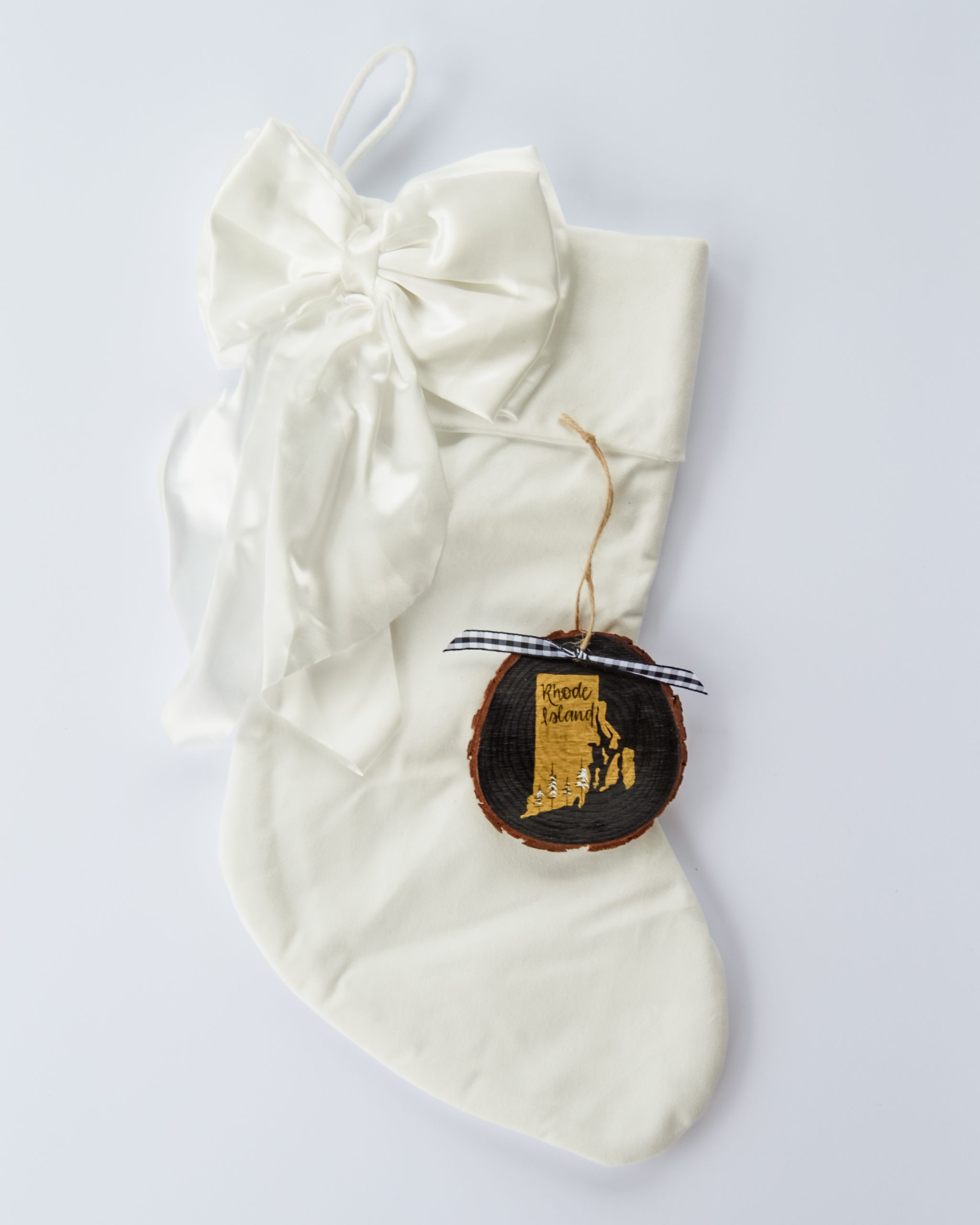 Hand-painted wood slice ornament of Rhode Island with a gold state silhouette, snow-covered trees, and the state name hand-lettered in black, tied with white buffalo plaid ribbon on a white velvet Christmas stocking