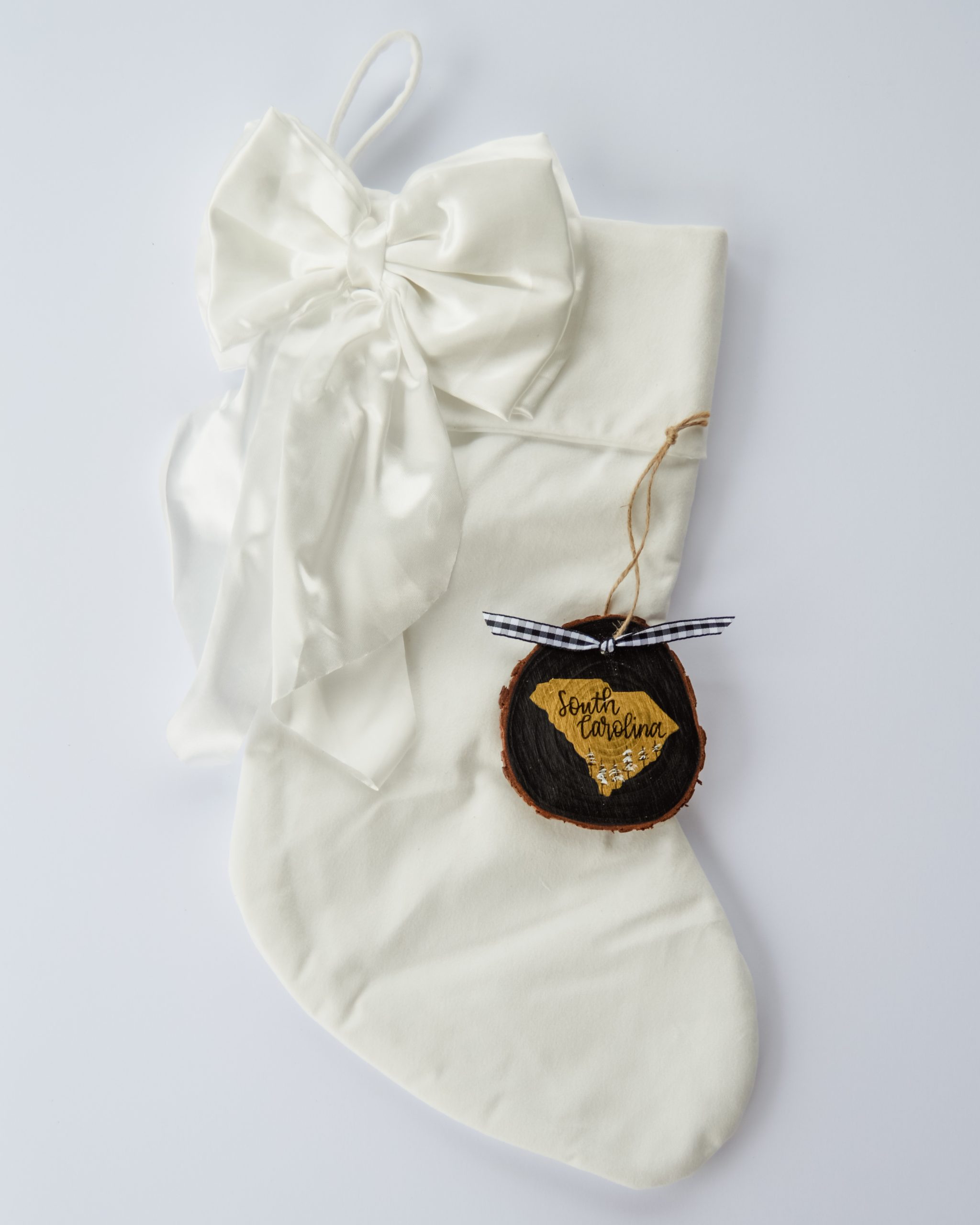 Hand-painted wood slice ornament of South Carolina with a gold state silhouette, snow-covered trees, and the state name hand-lettered in black, tied with white buffalo plaid ribbon on a white velvet Christmas stocking