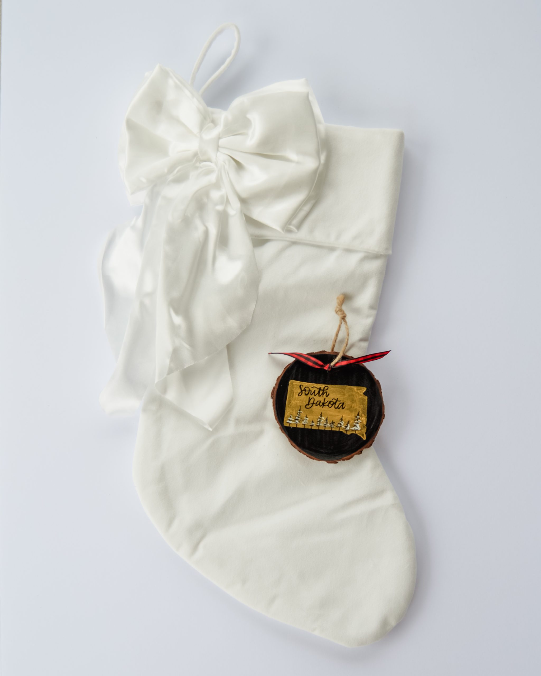 Hand-painted wood slice ornament of South Dakota with a gold state silhouette, snow-covered trees, and the state name hand-lettered in black, tied with red buffalo plaid ribbon on a white velvet Christmas stocking
