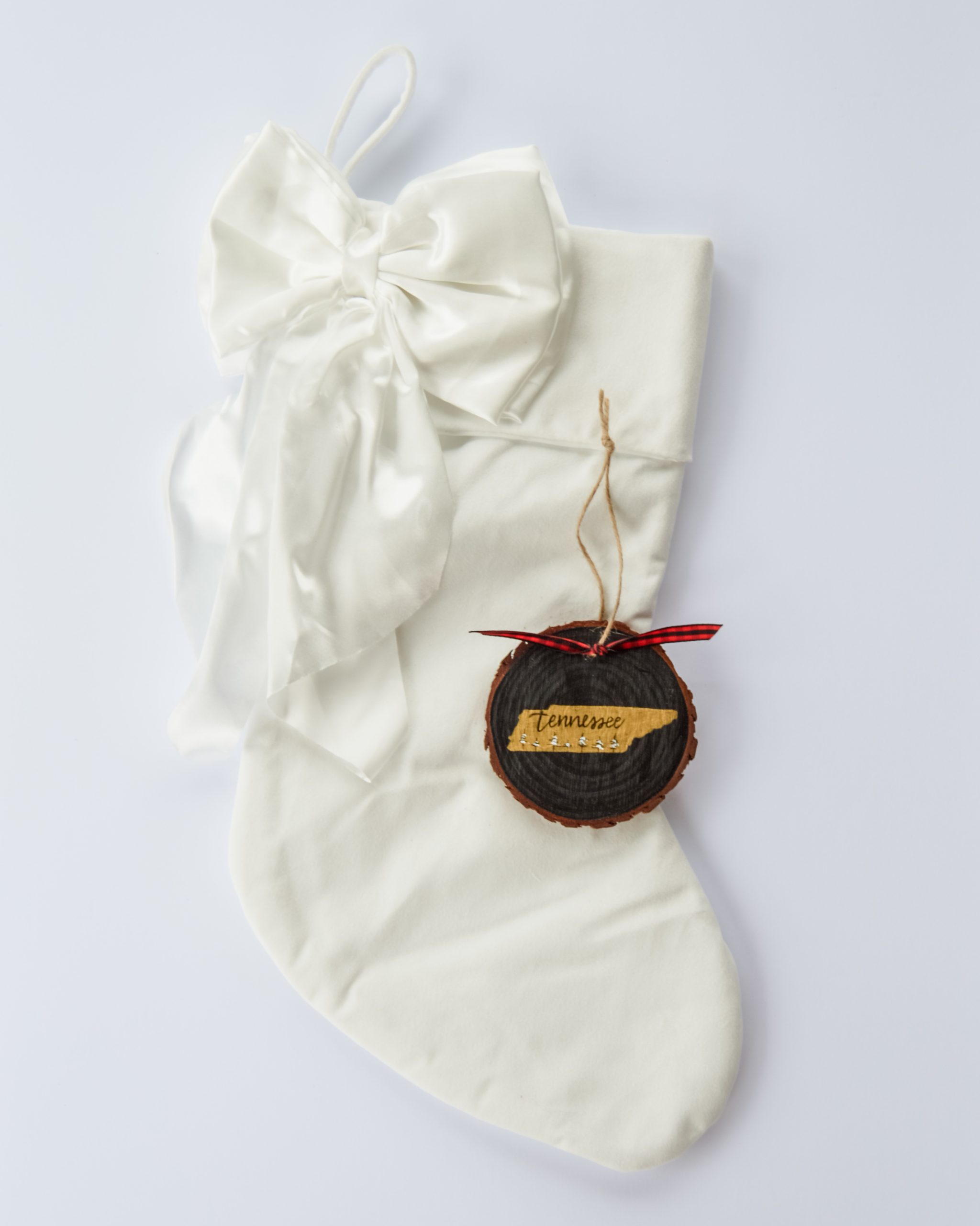 Hand-painted wood slice ornament of Tennessee with a gold state silhouette, snow-covered trees, and the state name hand-lettered in black, tied with red buffalo plaid ribbon on a white velvet Christmas stocking