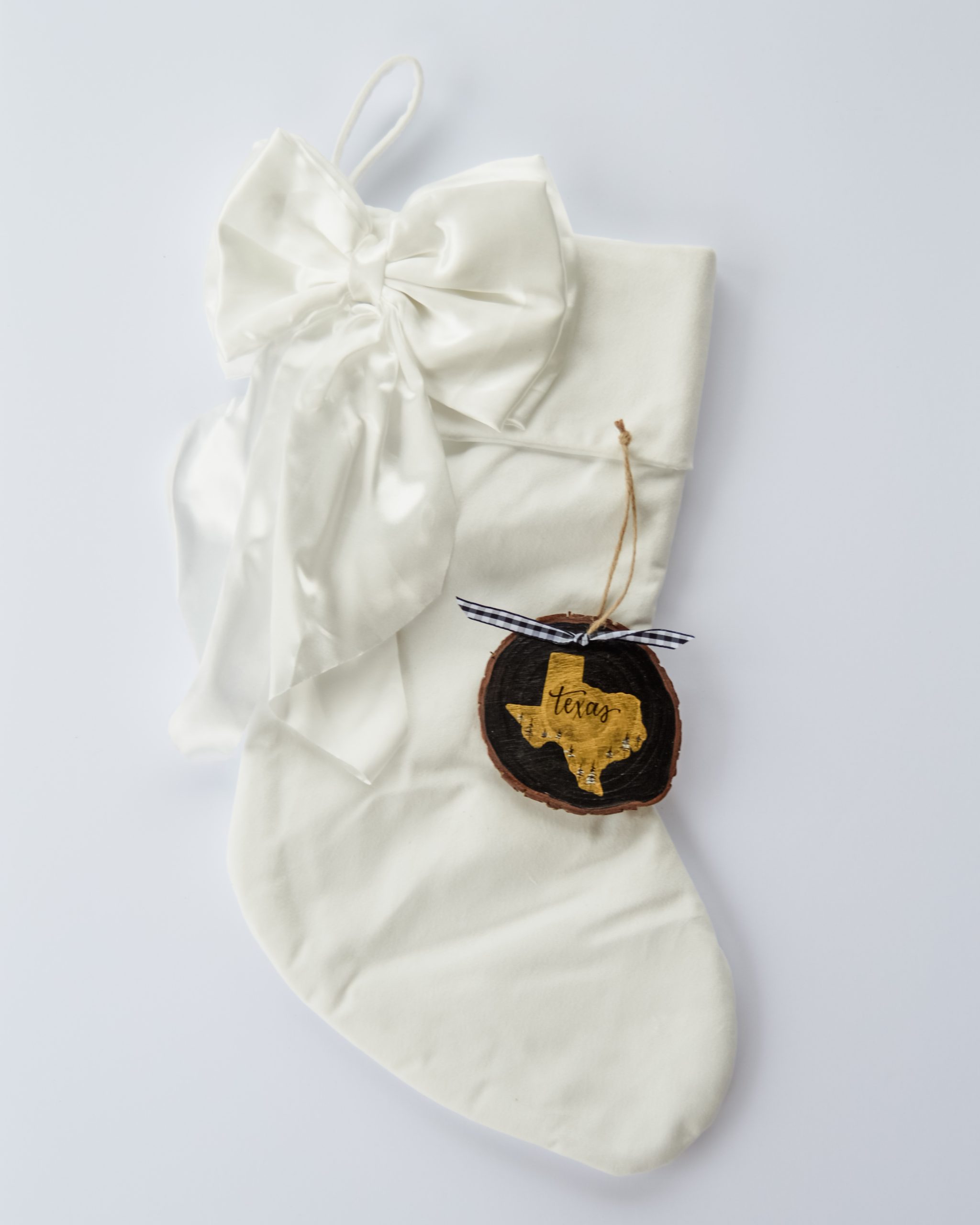 Hand-painted wood slice ornament of Texas with a gold state silhouette, snow-covered trees, and the state name hand-lettered in black, tied with white buffalo plaid ribbon on a white velvet Christmas stocking