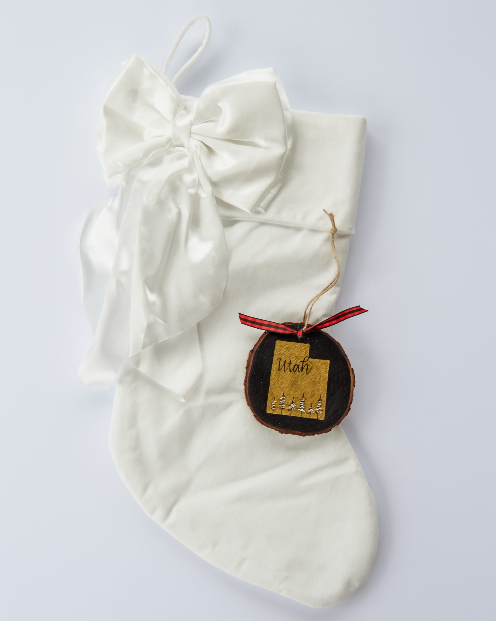 Hand-painted wood slice ornament of Utah with a gold state silhouette, snow-covered trees, and the state name hand-lettered in black, tied with red buffalo plaid ribbon on a white velvet Christmas stocking