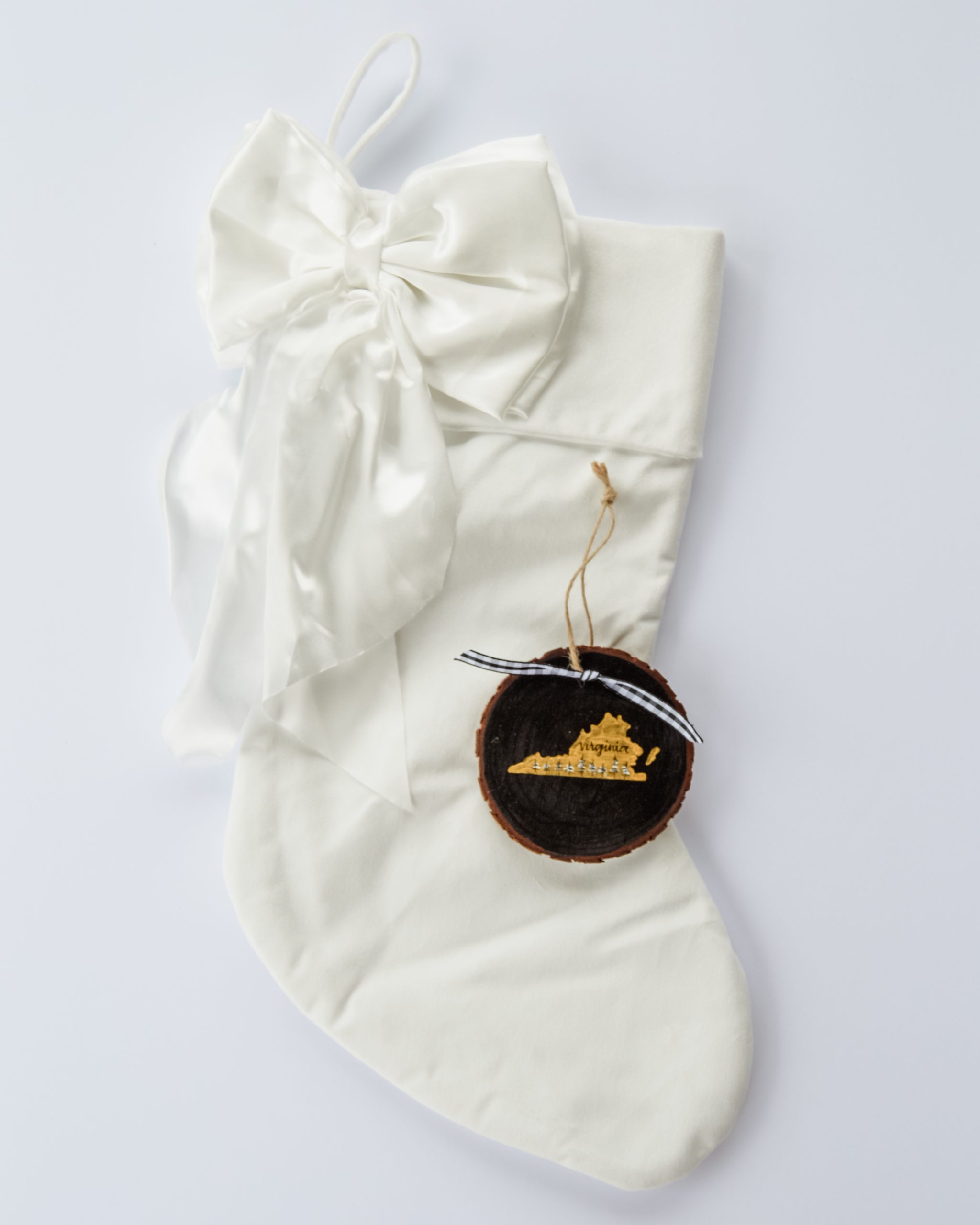 Hand-painted wood slice ornament of Virginia with a gold state silhouette, snow-covered trees, and the state name hand-lettered in black, tied with white buffalo plaid ribbon on a white velvet Christmas stocking