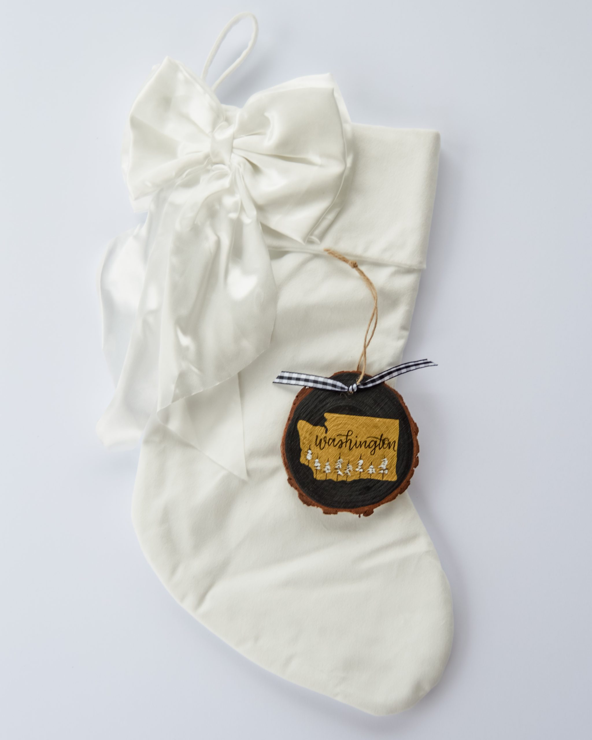 Hand-painted wood slice ornament of Washington with a gold state silhouette, snow-covered trees, and the state name hand-lettered in black, tied with white buffalo plaid ribbon on a white velvet Christmas stocking