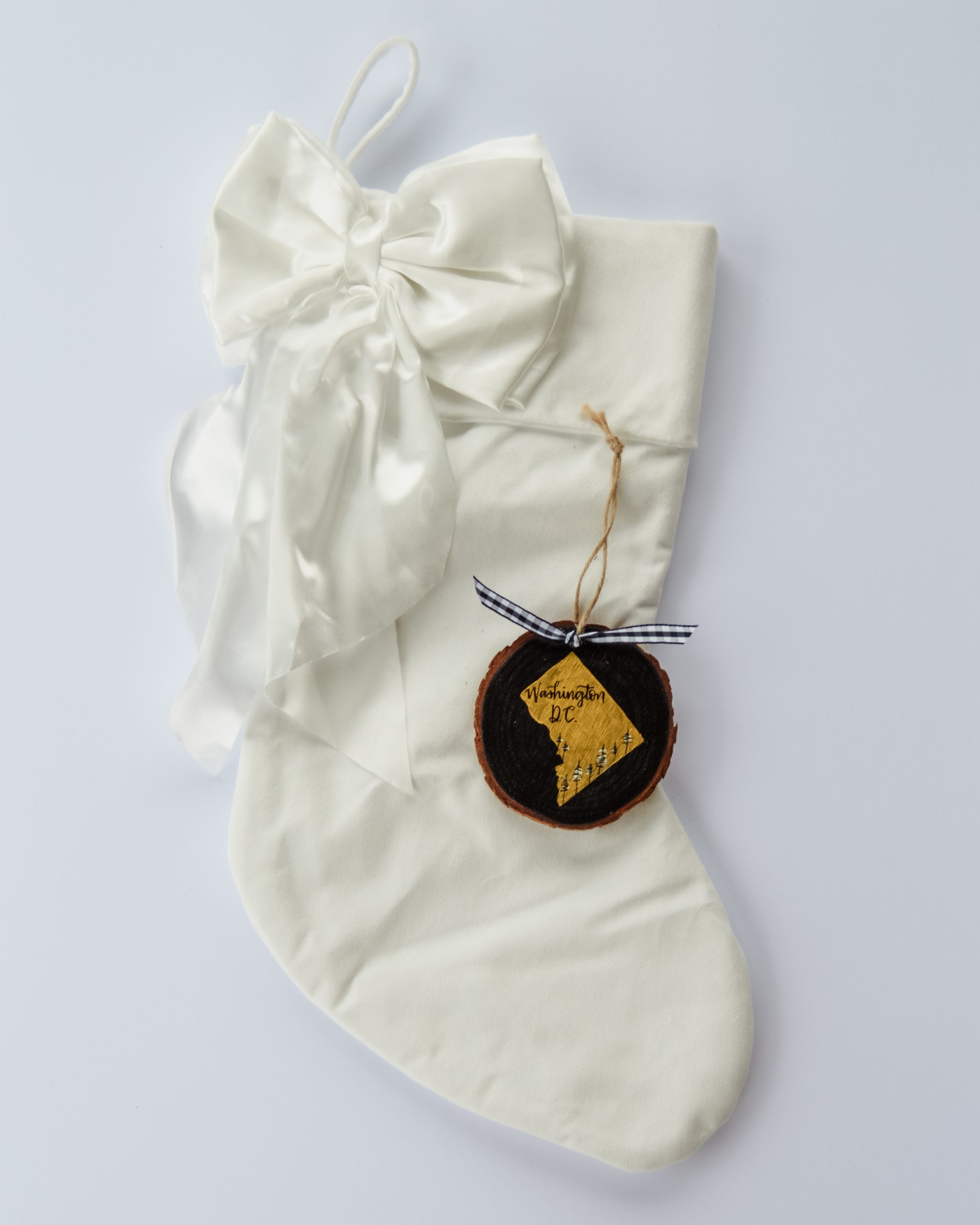 Hand-painted wood slice ornament of Washington DC with a gold state silhouette, snow-covered trees, and the state name hand-lettered in black, tied with white buffalo plaid ribbon on a white velvet Christmas stocking