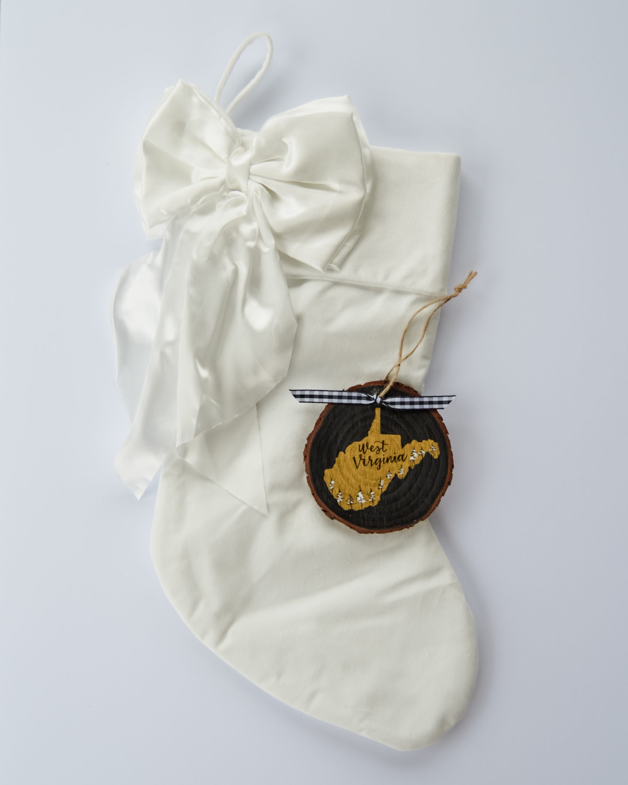 Hand-painted wood slice ornament of West Virginia with a gold state silhouette, snow-covered trees, and the state name hand-lettered in black, tied with white buffalo plaid ribbon on a white velvet Christmas stocking