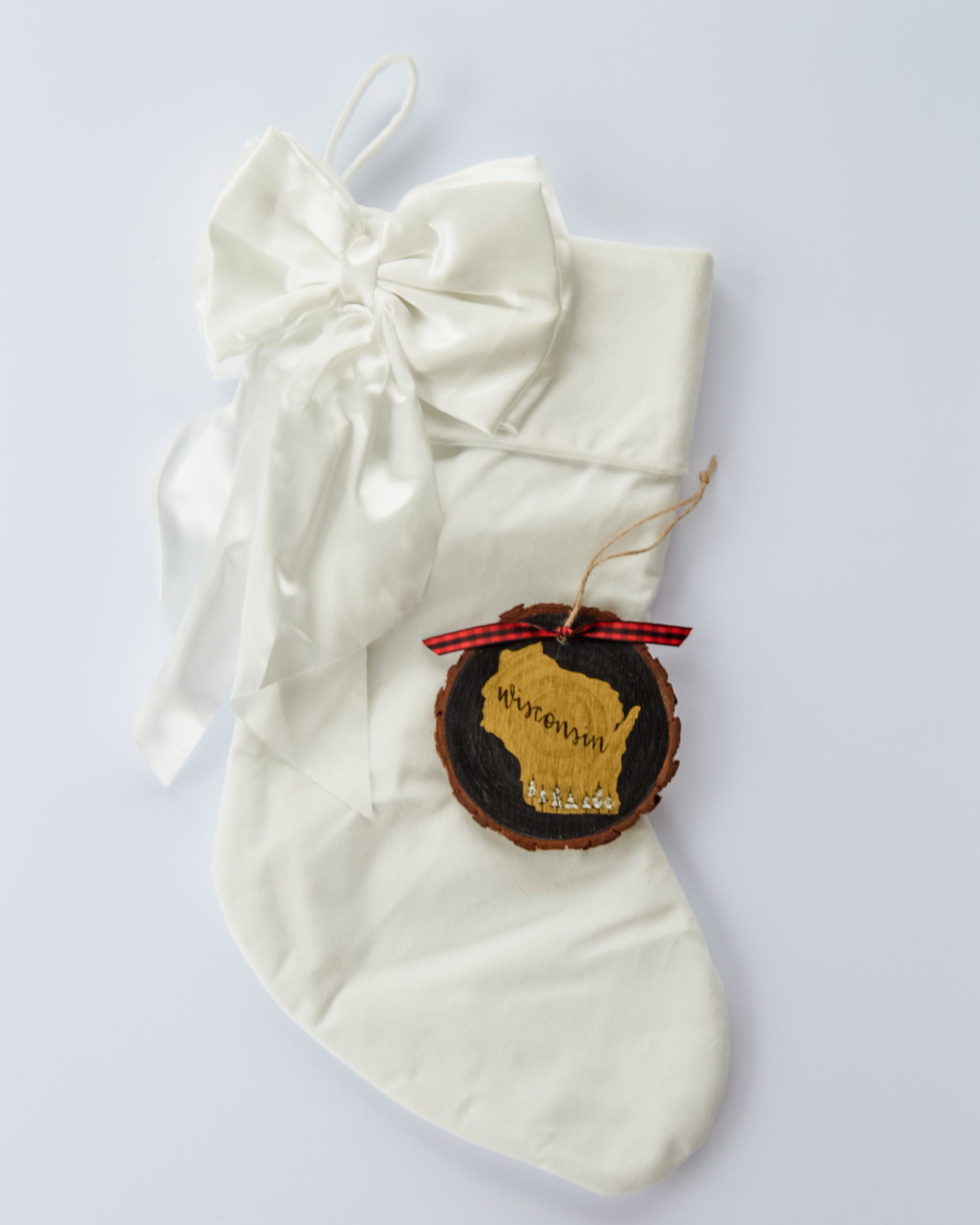 Hand-painted wood slice ornament of Wisconsin with a gold state silhouette, snow-covered trees, and the state name hand-lettered in black, tied with red buffalo plaid ribbon on a white velvet Christmas stocking