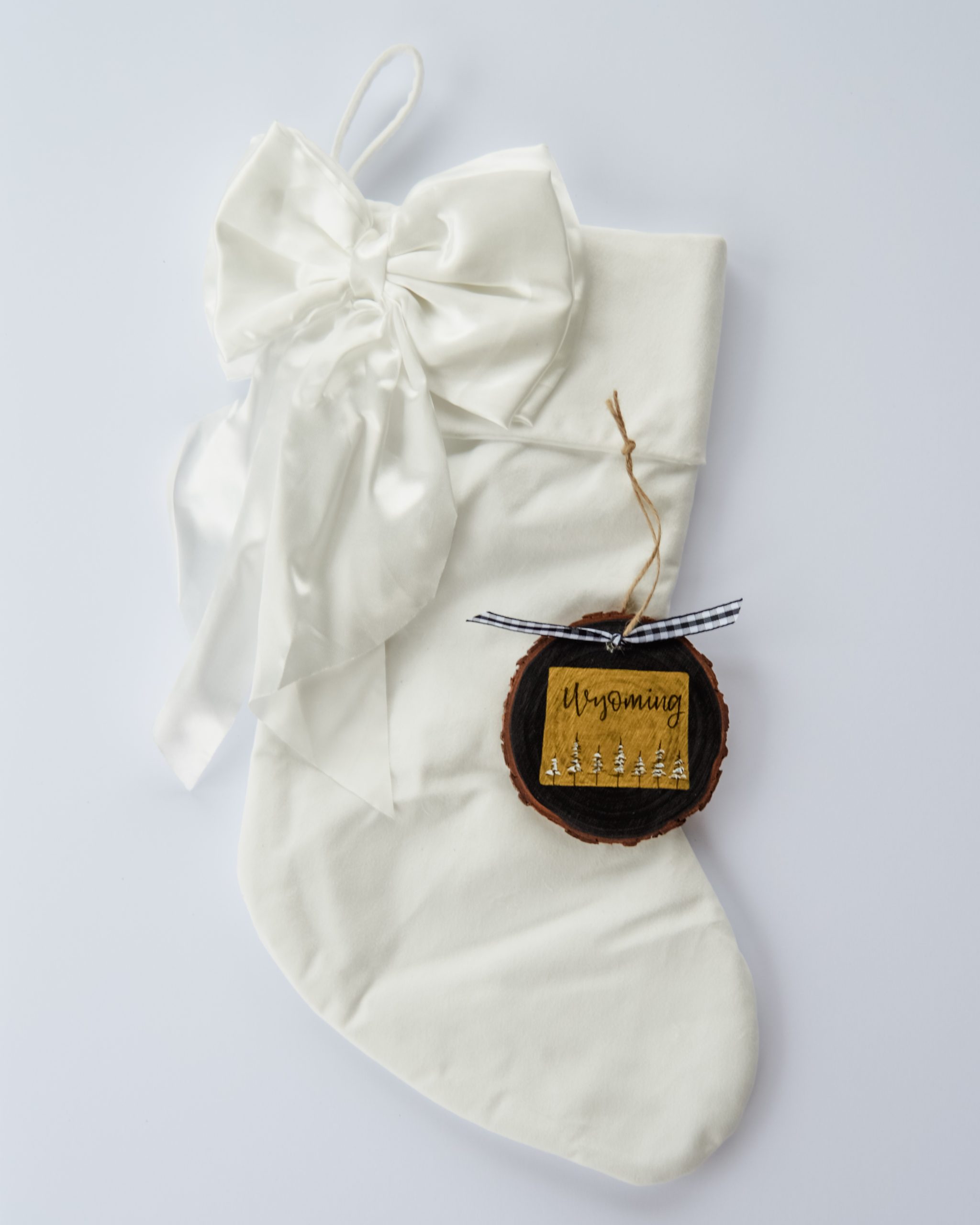 Hand-painted wood slice ornament of Wyoming with a gold state silhouette, snow-covered trees, and the state name hand-lettered in black, tied with white buffalo plaid ribbon on a white velvet Christmas stocking