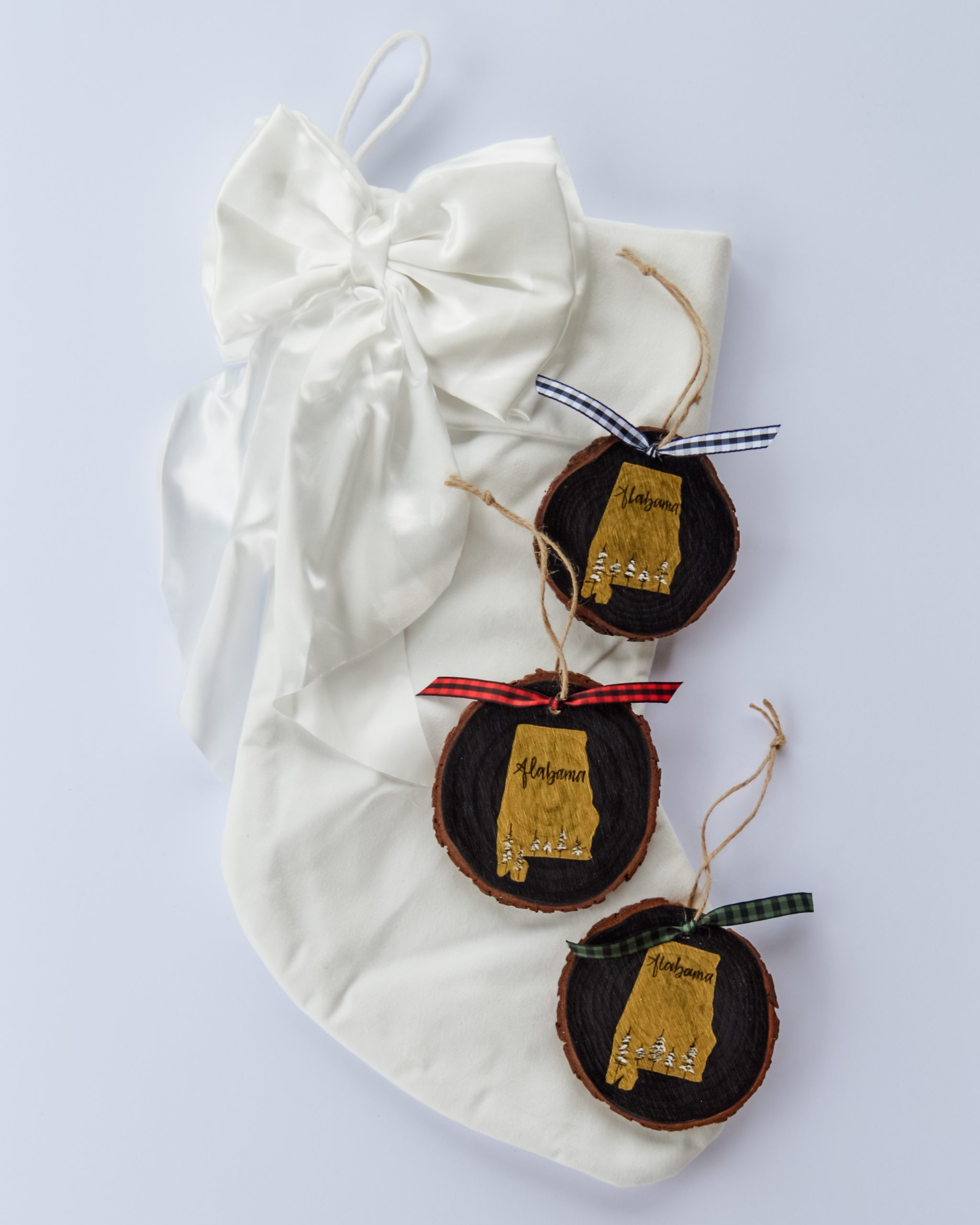 Hand-painted wood slice ornaments of Alabama with a gold state silhouette, snow-covered trees, and the state name hand-lettered in black, tied with white, red, and hunter green buffalo plaid ribbons on a white velvet Christmas stocking