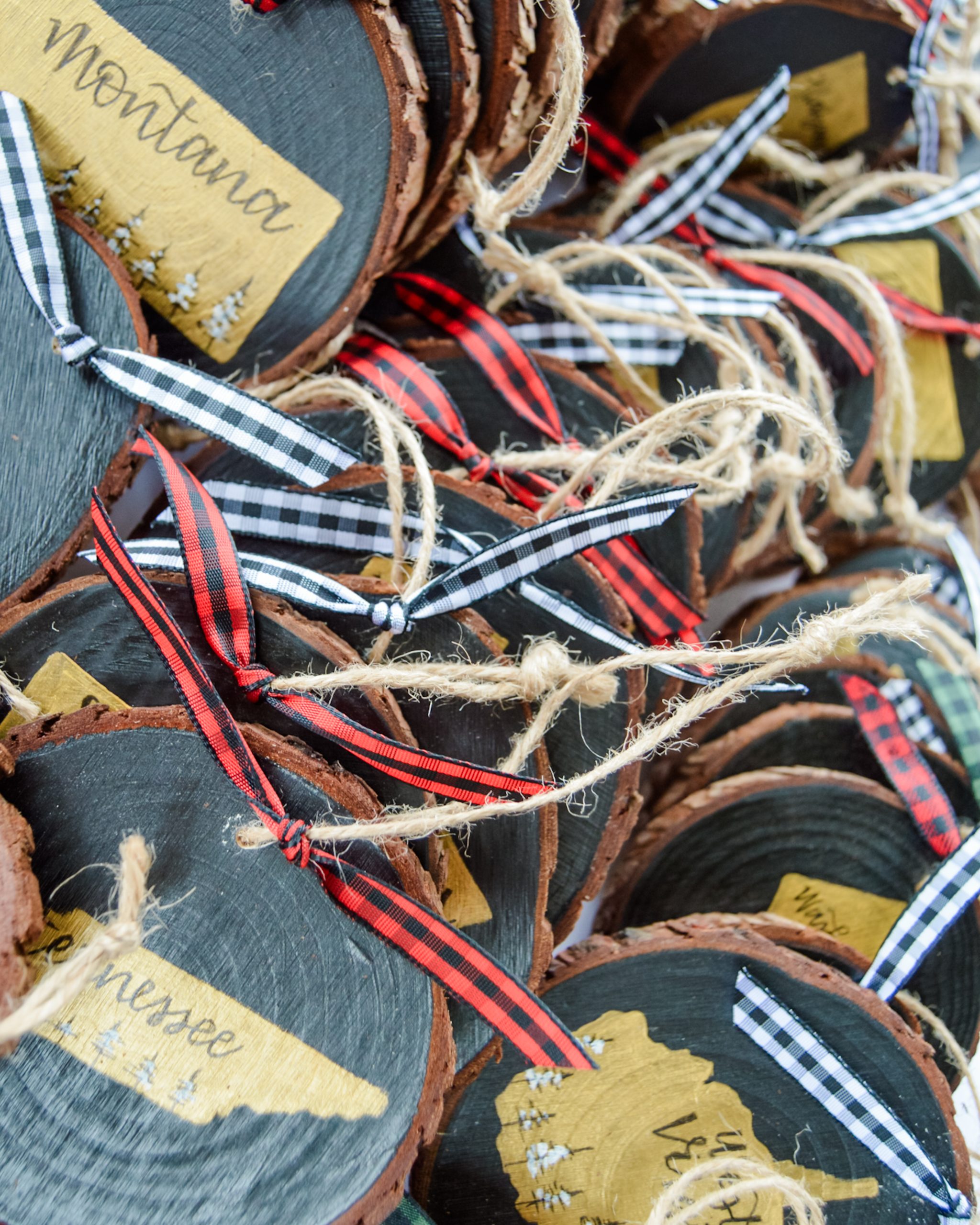 A pile of hand-painted wood slice Christmas ornaments with gold state silhouettes on black backgrounds, including Montana, Tennessee, and West Virginia, featuring snow-covered trees, hand-lettered state names, jute twine hangers, and red and white buffalo plaid ribbons