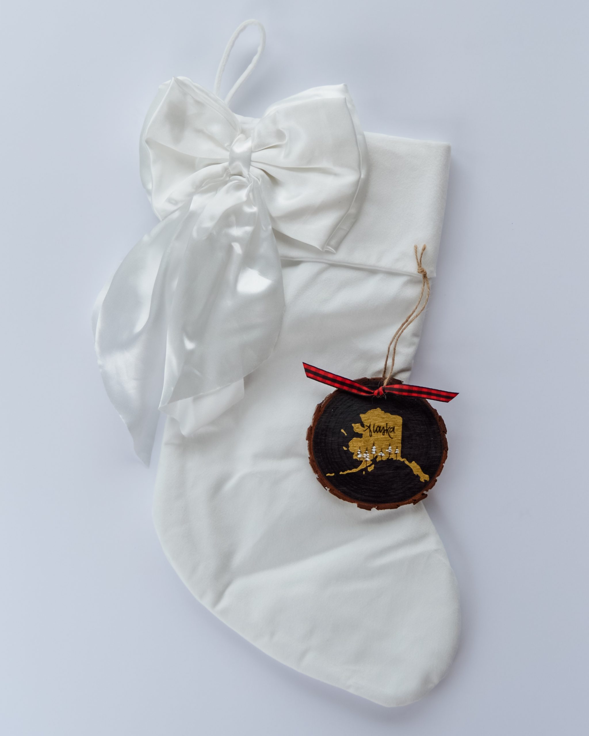 Hand-painted wood slice ornament of Alaska with a gold state silhouette, snow-covered trees, and the state name hand-lettered in black, tied with red buffalo plaid ribbon on a white velvet Christmas stocking