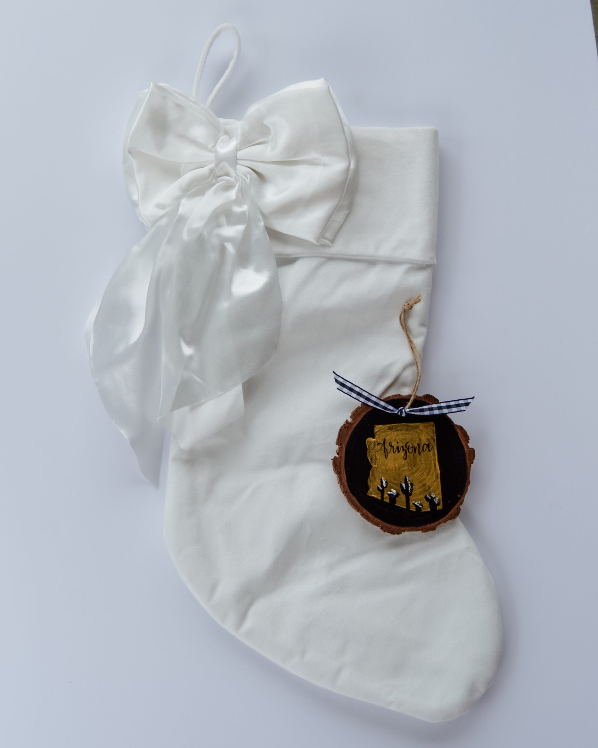 Hand-painted wood slice ornament of Arizona with a gold state silhouette, snow-covered cacti, and the state name hand-lettered in black, tied with white buffalo plaid ribbon on a white velvet Christmas stocking