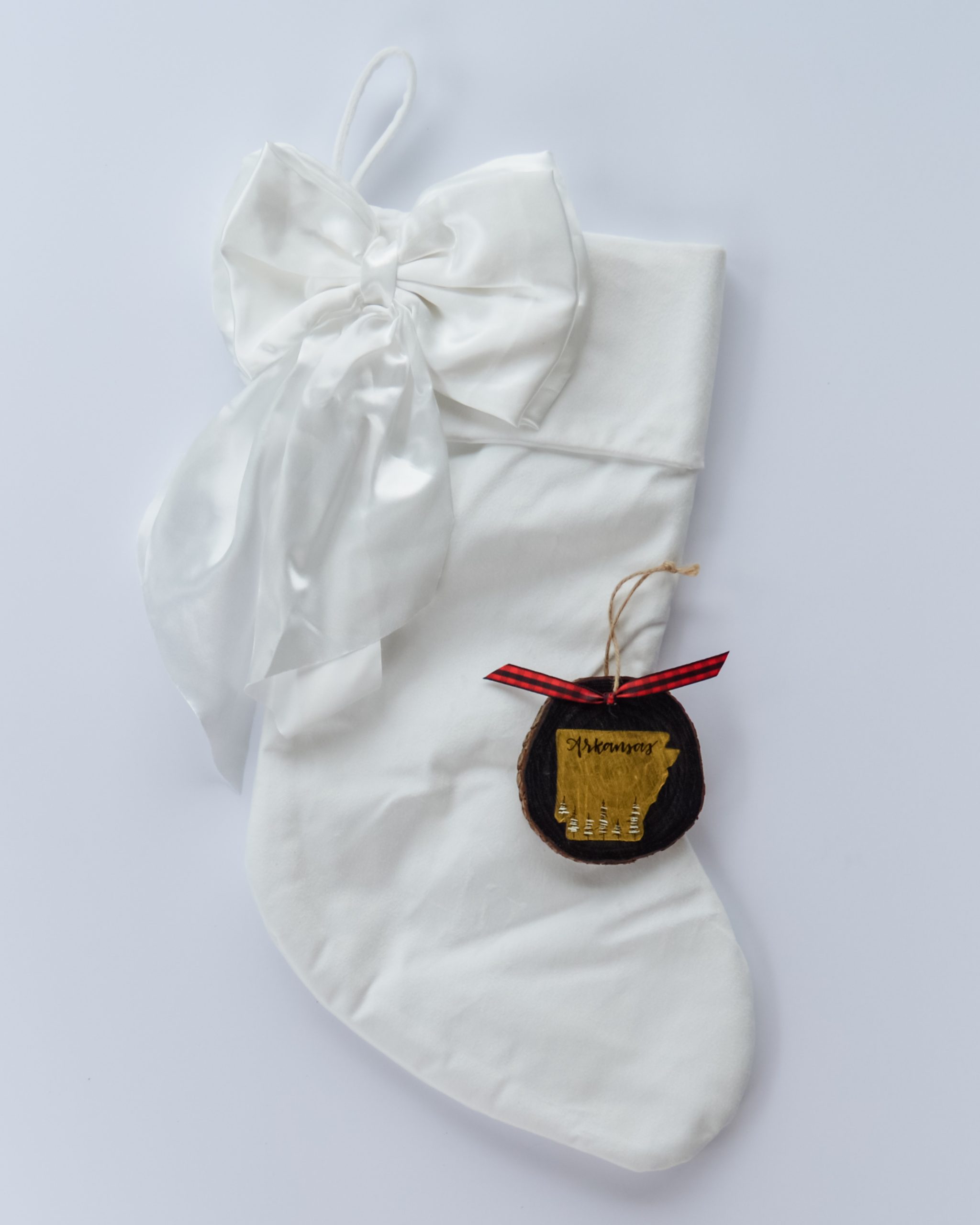 Hand-painted wood slice ornament of Arkansas with a gold state silhouette, snow-covered trees, and the state name hand-lettered in black, tied with red buffalo plaid ribbon on a white velvet Christmas stocking