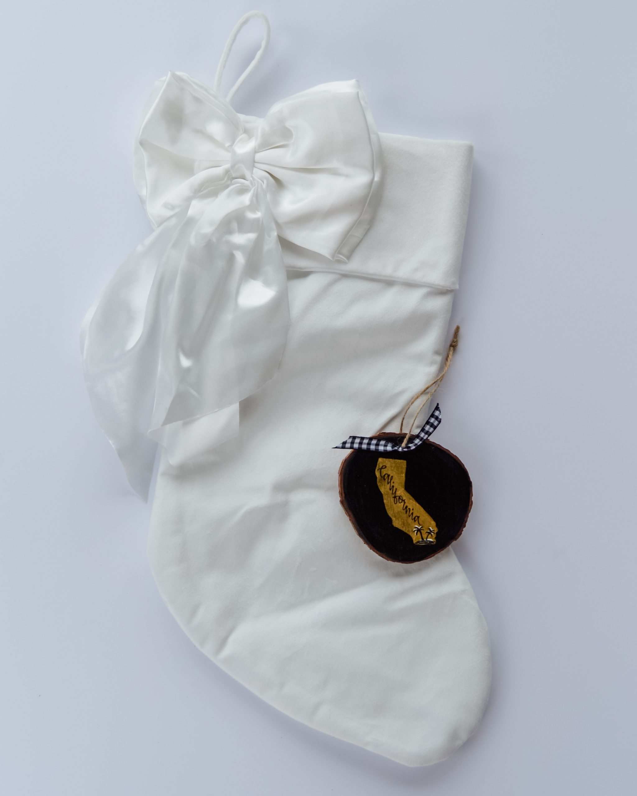 Hand-painted wood slice ornament of California with a gold state silhouette, snow-covered palm trees, and the state name hand-lettered in black, tied with white buffalo plaid ribbon on a white velvet Christmas stocking