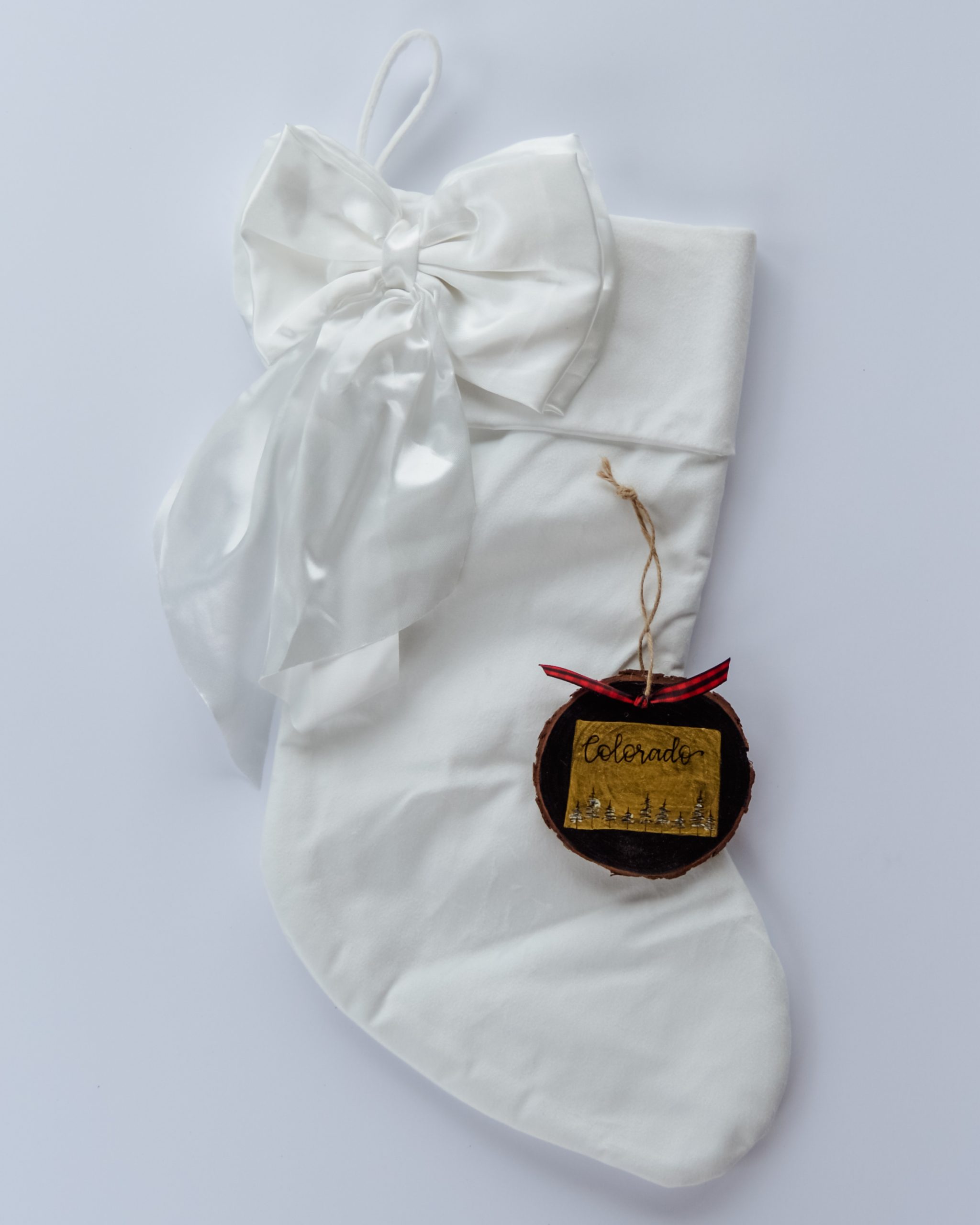 Hand-painted wood slice ornament of Colorado with a gold state silhouette, snow-covered trees, and the state name hand-lettered in black, tied with red buffalo plaid ribbon on a white velvet Christmas stocking
