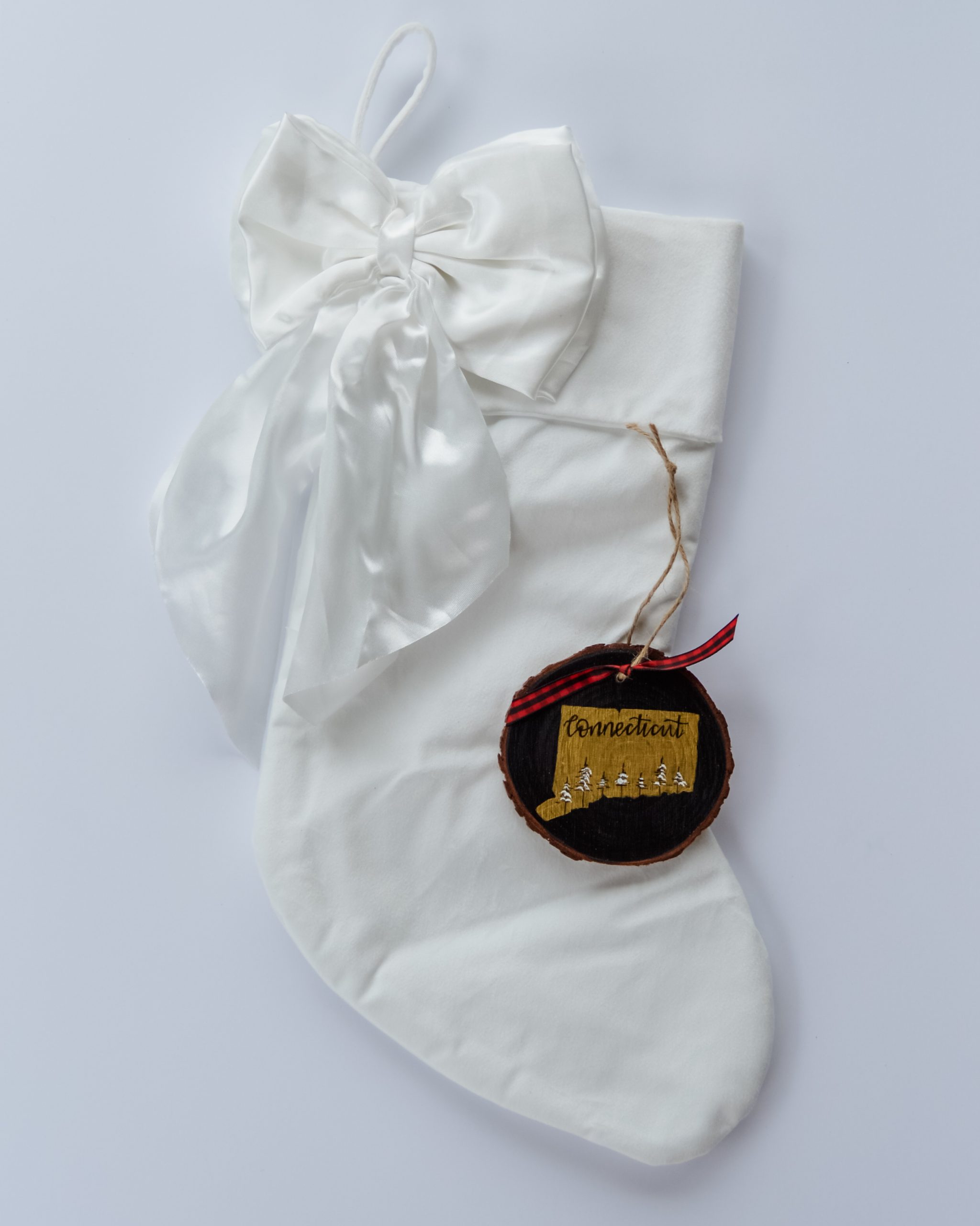 Hand-painted wood slice ornament of Connecticut with a gold state silhouette, snow-covered trees, and the state name hand-lettered in black, tied with red buffalo plaid ribbon on a white velvet Christmas stocking
