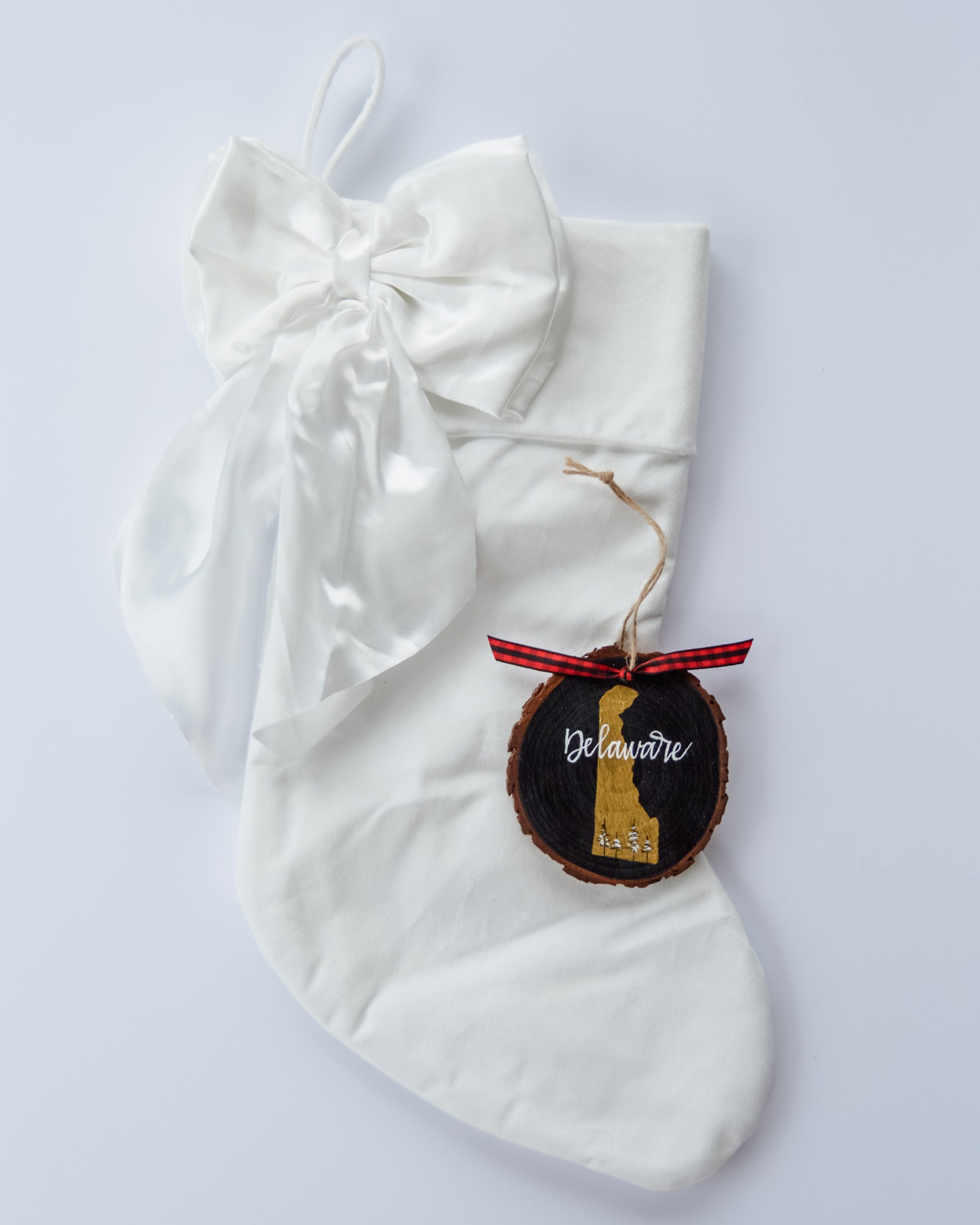 Hand-painted wood slice ornament of Delaware with a gold state silhouette, snow-covered trees, and the state name hand-lettered in white, tied with white buffalo plaid ribbon on a white velvet Christmas stocking