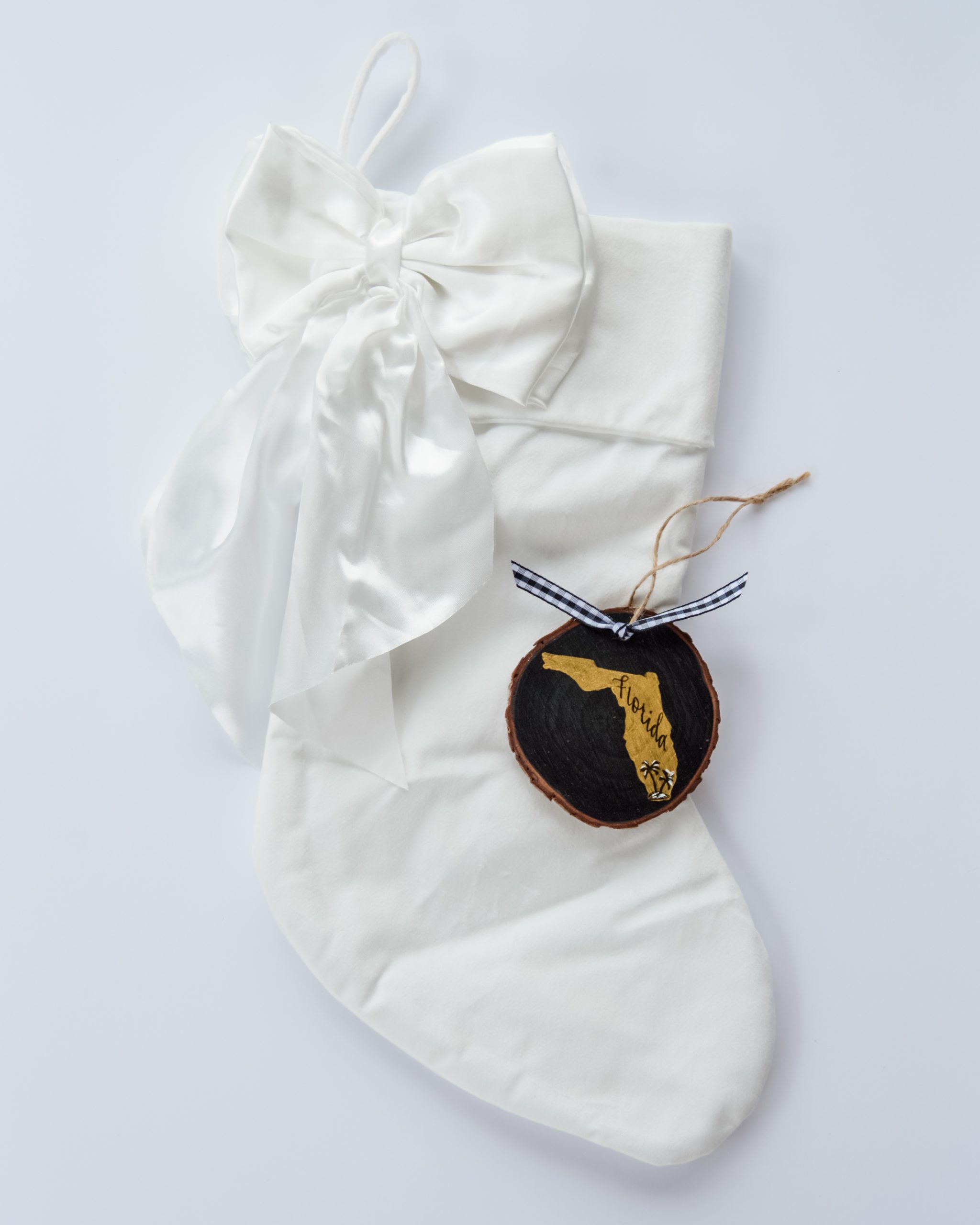 Hand-painted wood slice ornament of Florida with a gold state silhouette, snow-covered palm trees, and the state name hand-lettered in black, tied with white buffalo plaid ribbon on a white velvet Christmas stocking