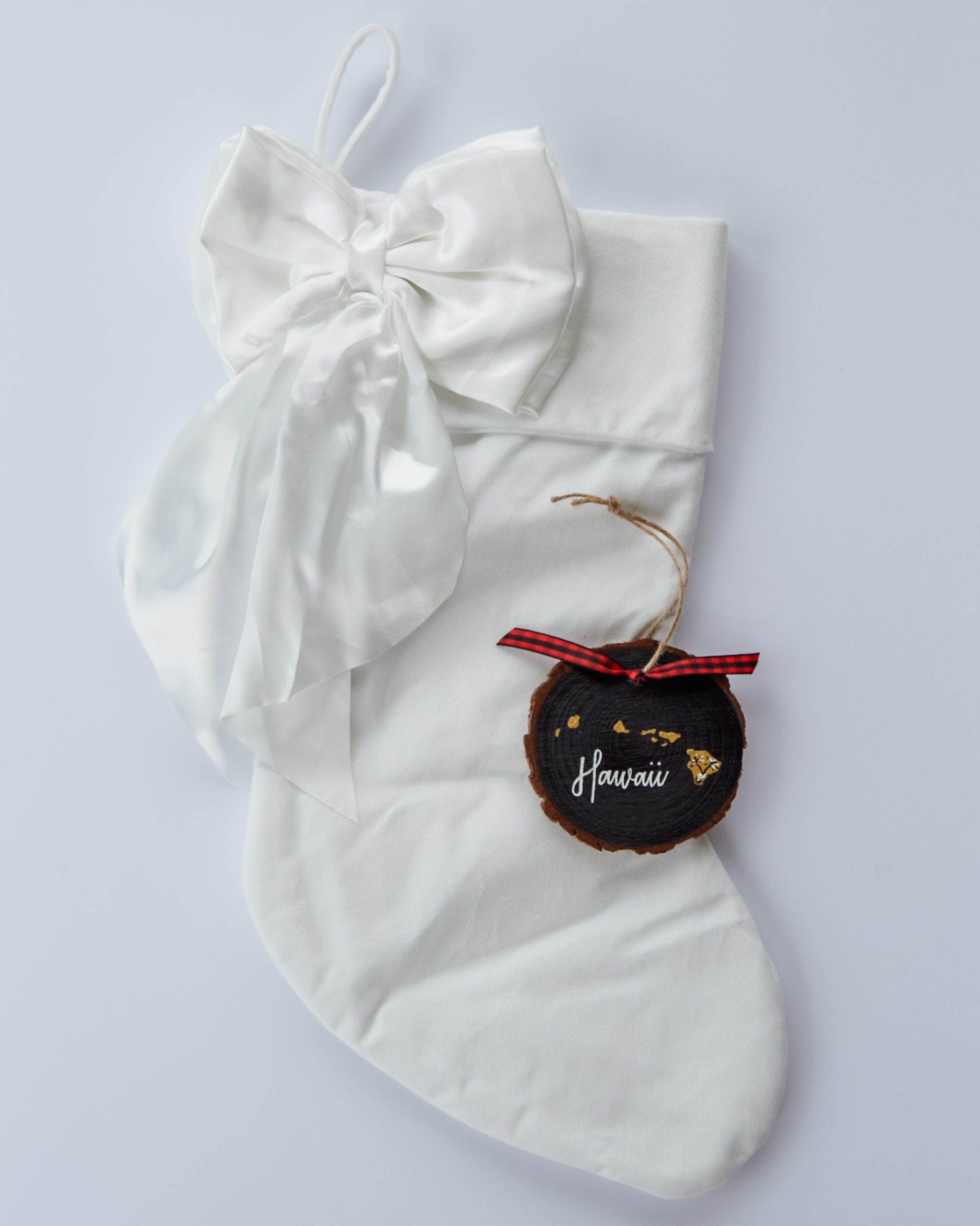 Hand-painted wood slice ornament of Hawaii with a gold state silhouette, snow-covered palm trees, and the state name hand-lettered in black, tied with red buffalo plaid ribbon on a white velvet Christmas stocking