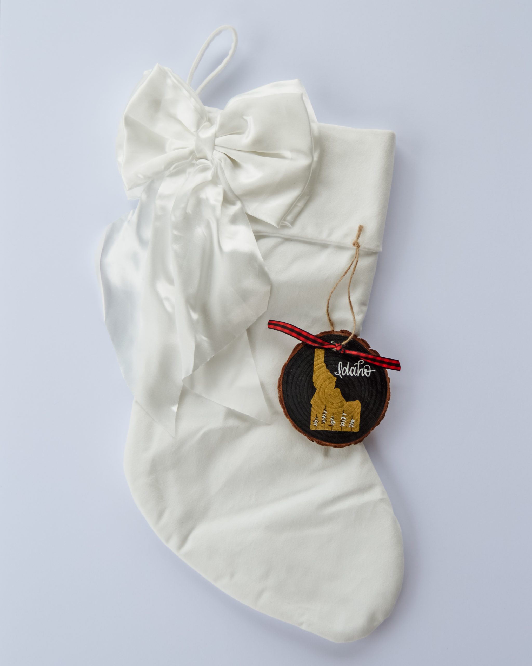 Hand-painted wood slice ornament of Idaho with a gold state silhouette, snow-covered trees, and the state name hand-lettered in white, tied with red buffalo plaid ribbon on a white velvet Christmas stocking