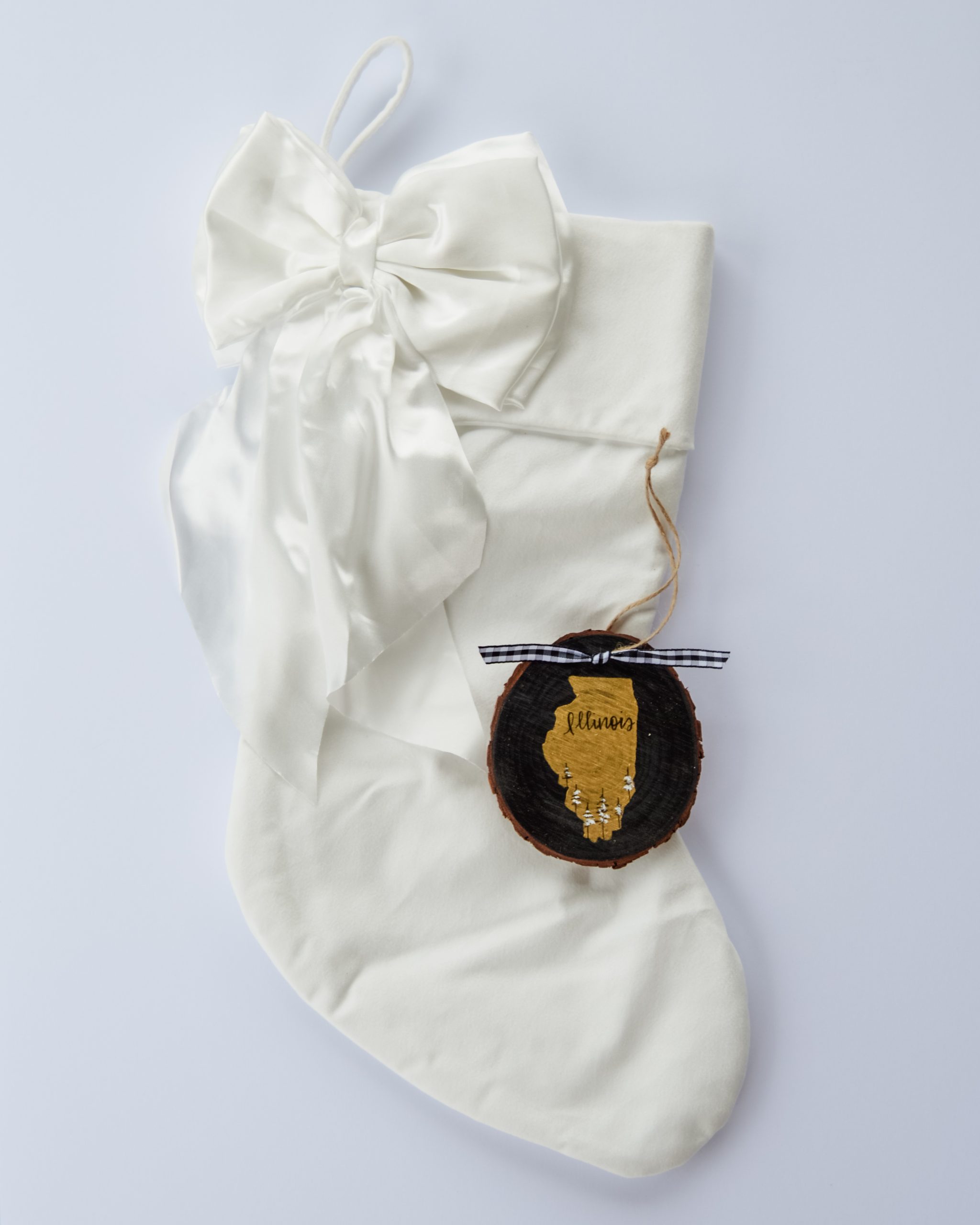 Hand-painted wood slice ornament of Illinois with a gold state silhouette, snow-covered trees, and the state name hand-lettered in black, tied with white buffalo plaid ribbon on a white velvet Christmas stocking