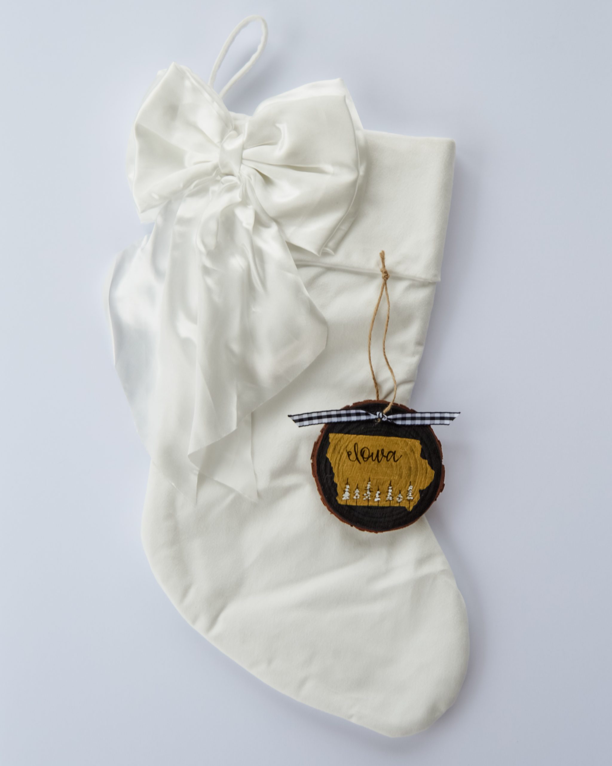 Hand-painted wood slice ornament of Iowa with a gold state silhouette, snow-covered trees, and the state name hand-lettered in black, tied with white buffalo plaid ribbon on a white velvet Christmas stocking