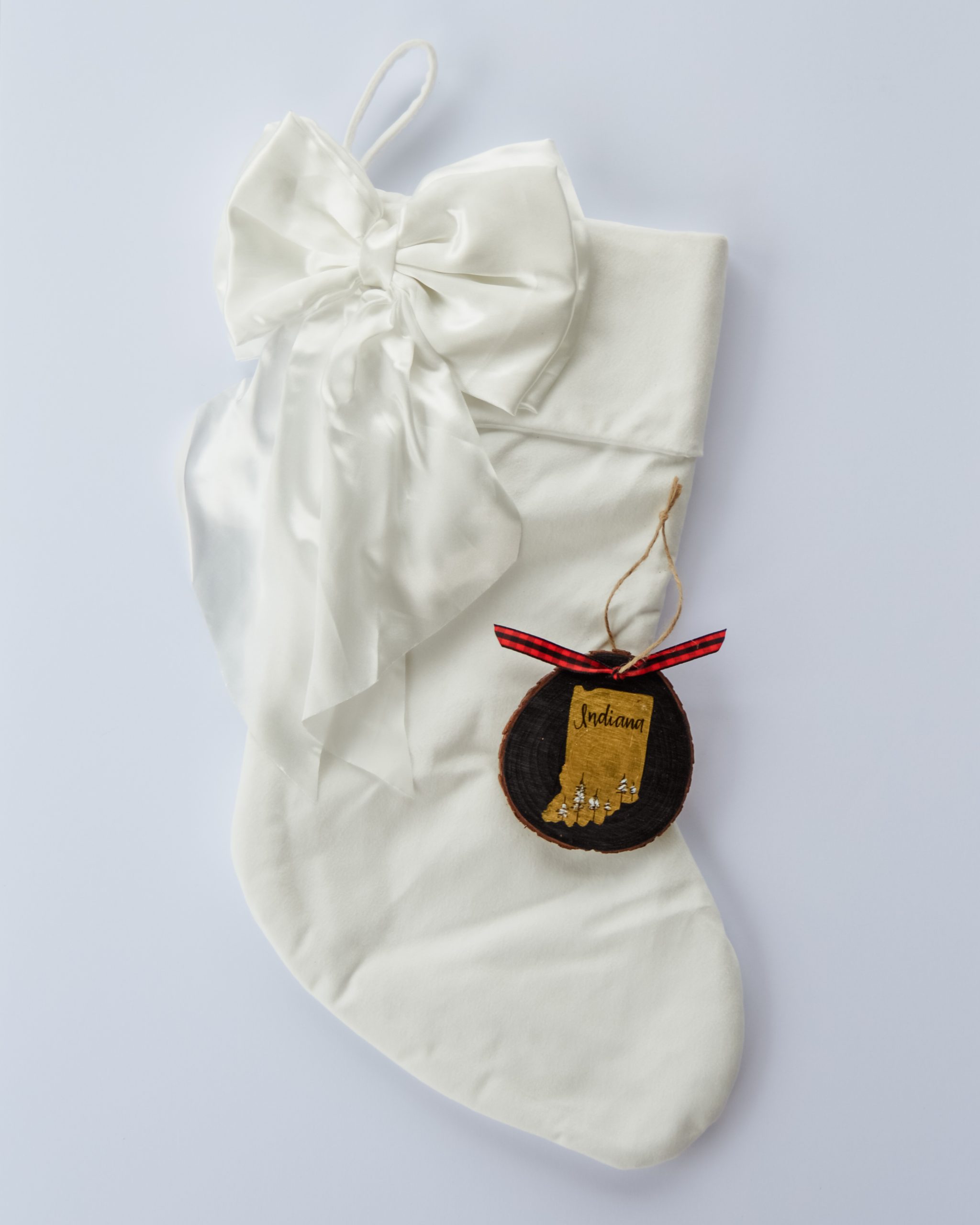 Hand-painted wood slice ornament of Indiana with a gold state silhouette, snow-covered trees, and the state name hand-lettered in black, tied with red buffalo plaid ribbon on a white velvet Christmas stocking