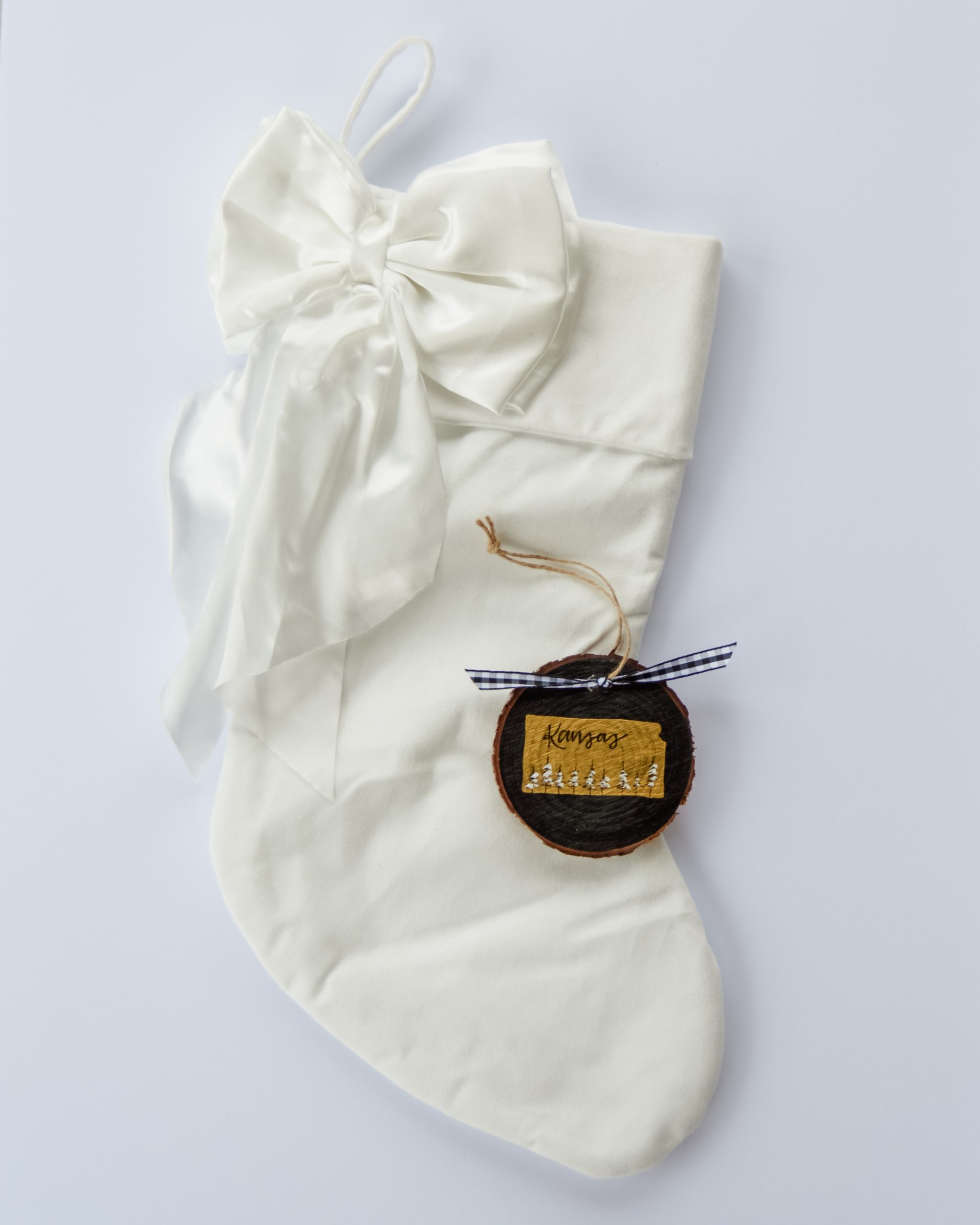 Hand-painted wood slice ornament of Kansas with a gold state silhouette, snow-covered trees, and the state name hand-lettered in black, tied with white buffalo plaid ribbon on a white velvet Christmas stocking