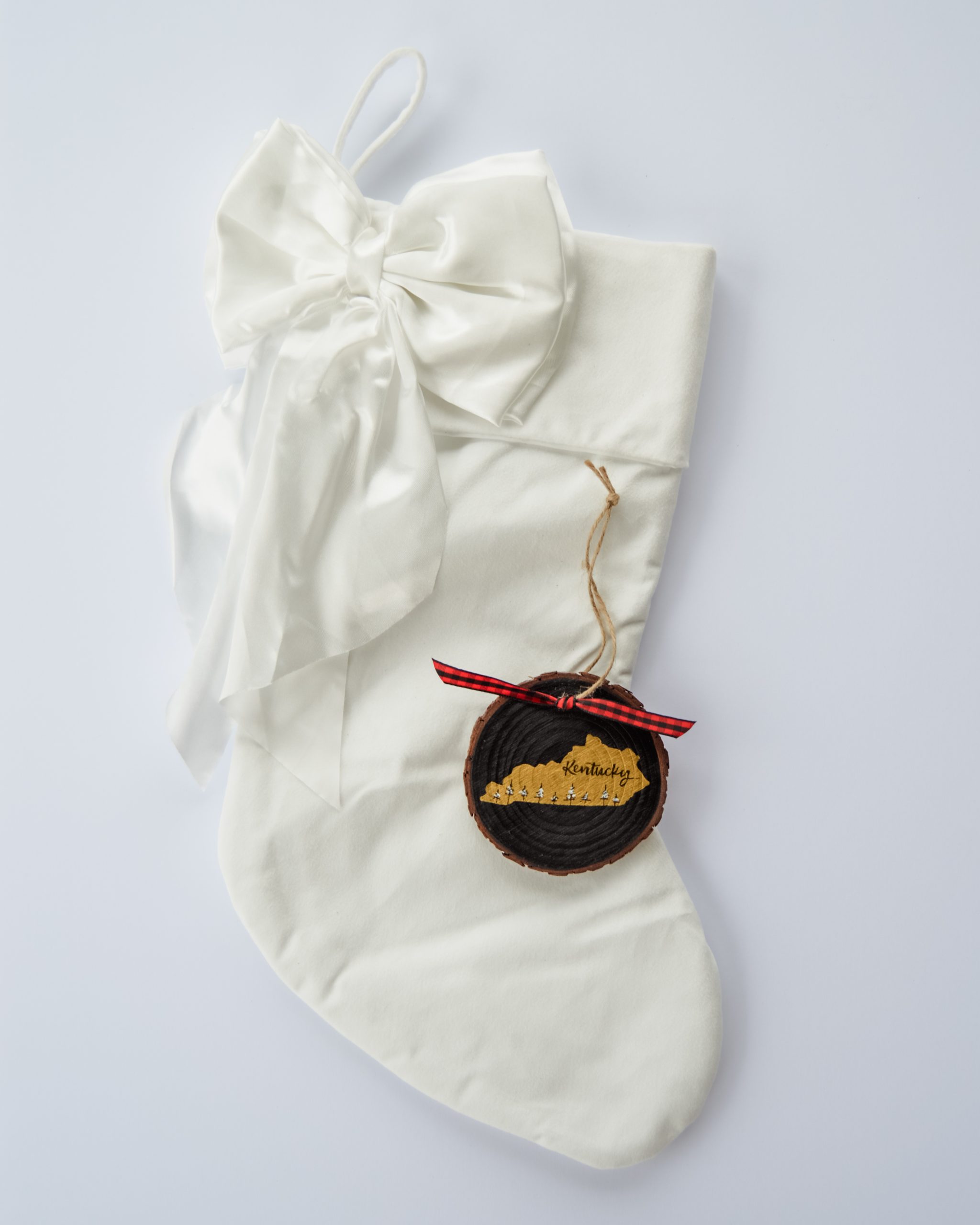 Hand-painted wood slice ornament of Kentucky with a gold state silhouette, snow-covered trees, and the state name hand-lettered in black, tied with red buffalo plaid ribbon on a white velvet Christmas stocking