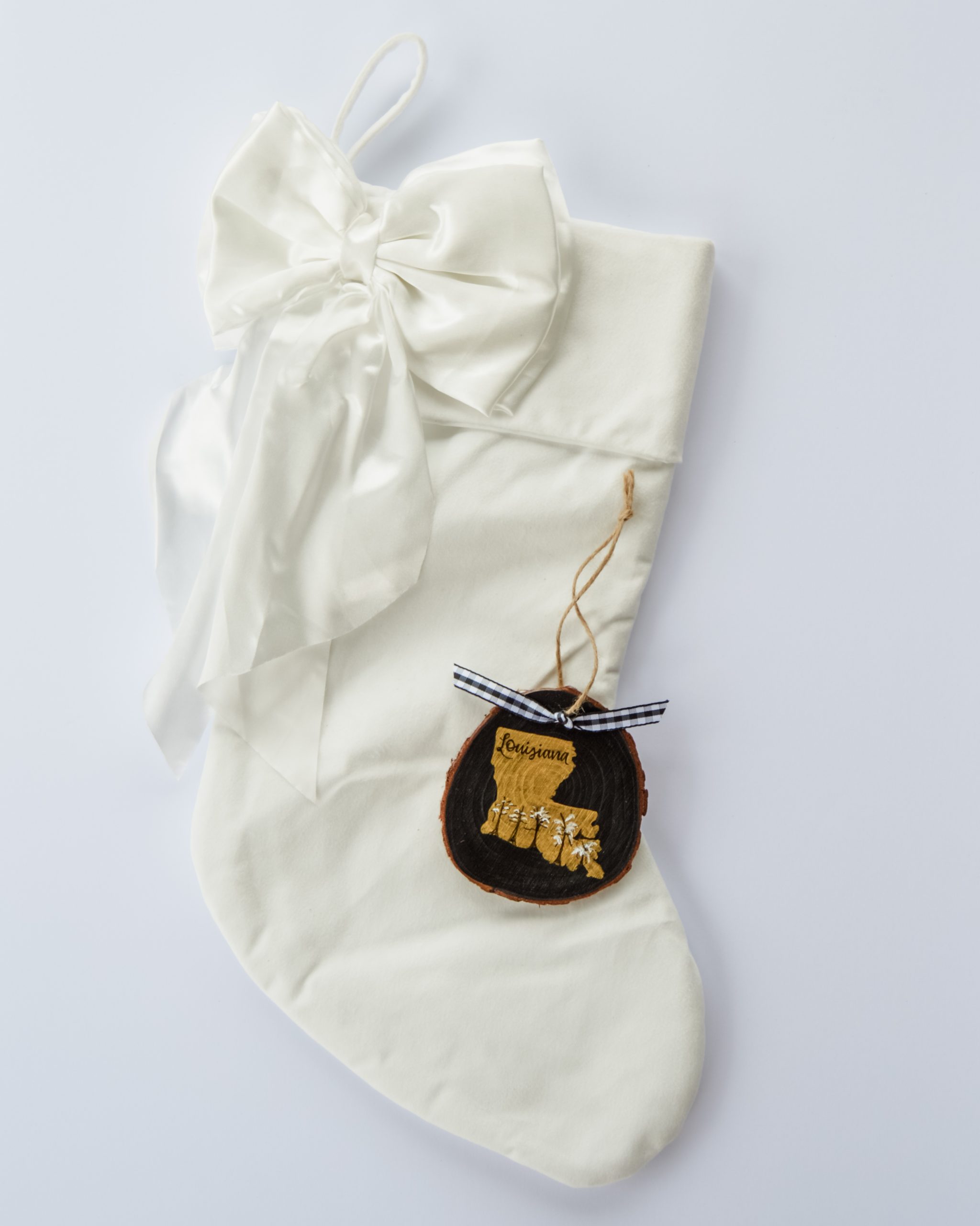 Hand-painted wood slice ornament of Louisiana with a gold state silhouette, snow-covered Cypress trees, and the state name hand-lettered in black, tied with white buffalo plaid ribbon on a white velvet Christmas stocking