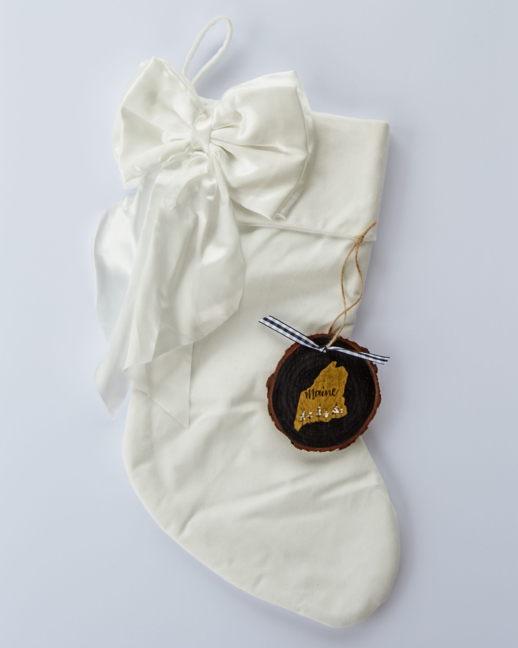 Hand-painted wood slice ornament of Maine with a gold state silhouette, snow-covered trees, and the state name hand-lettered in black, tied with white buffalo plaid ribbon on a white velvet Christmas stocking