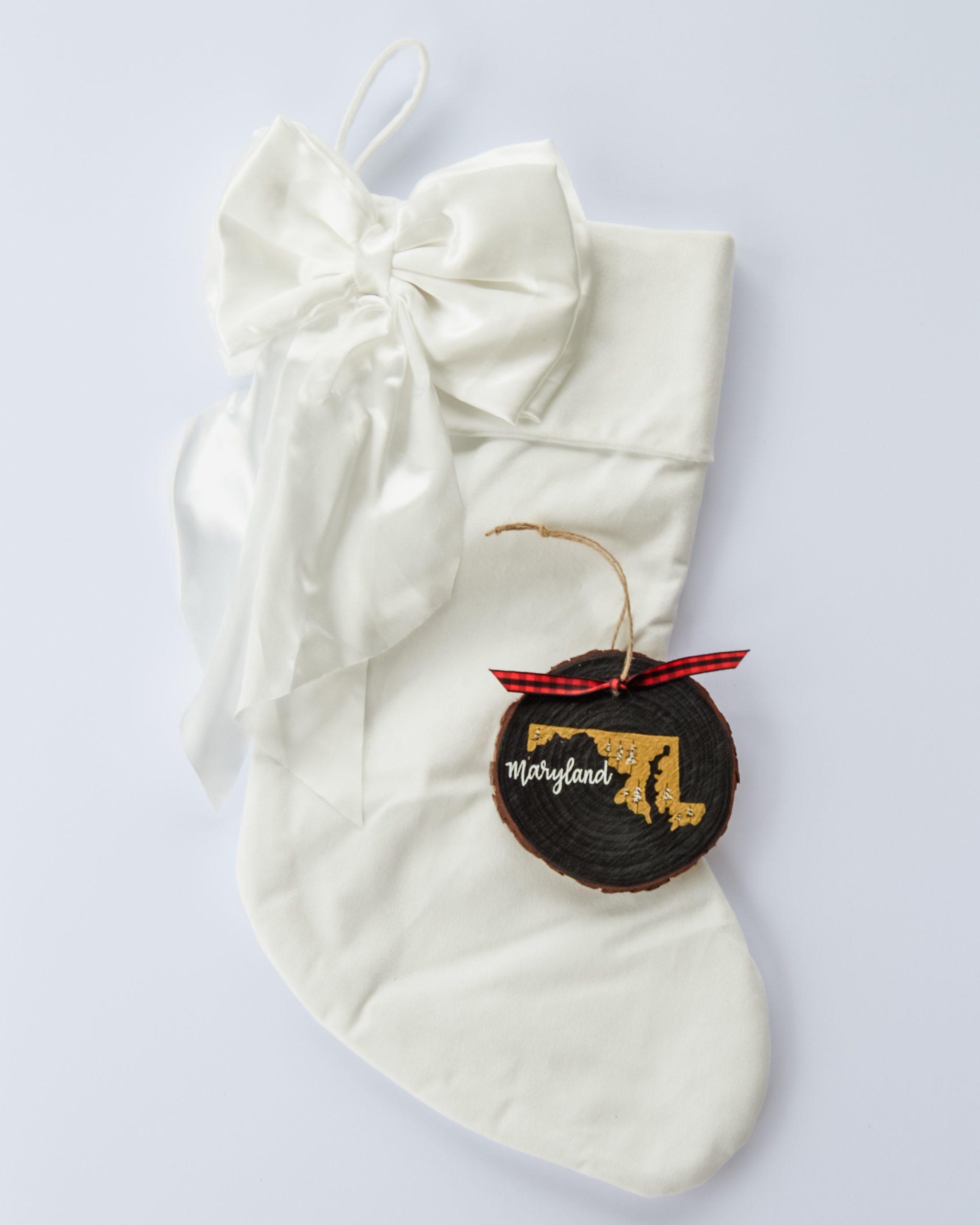 Hand-painted wood slice ornament of Maryland with a gold state silhouette, snow-covered trees, and the state name hand-lettered in white, tied with red buffalo plaid ribbon on a white velvet Christmas stocking