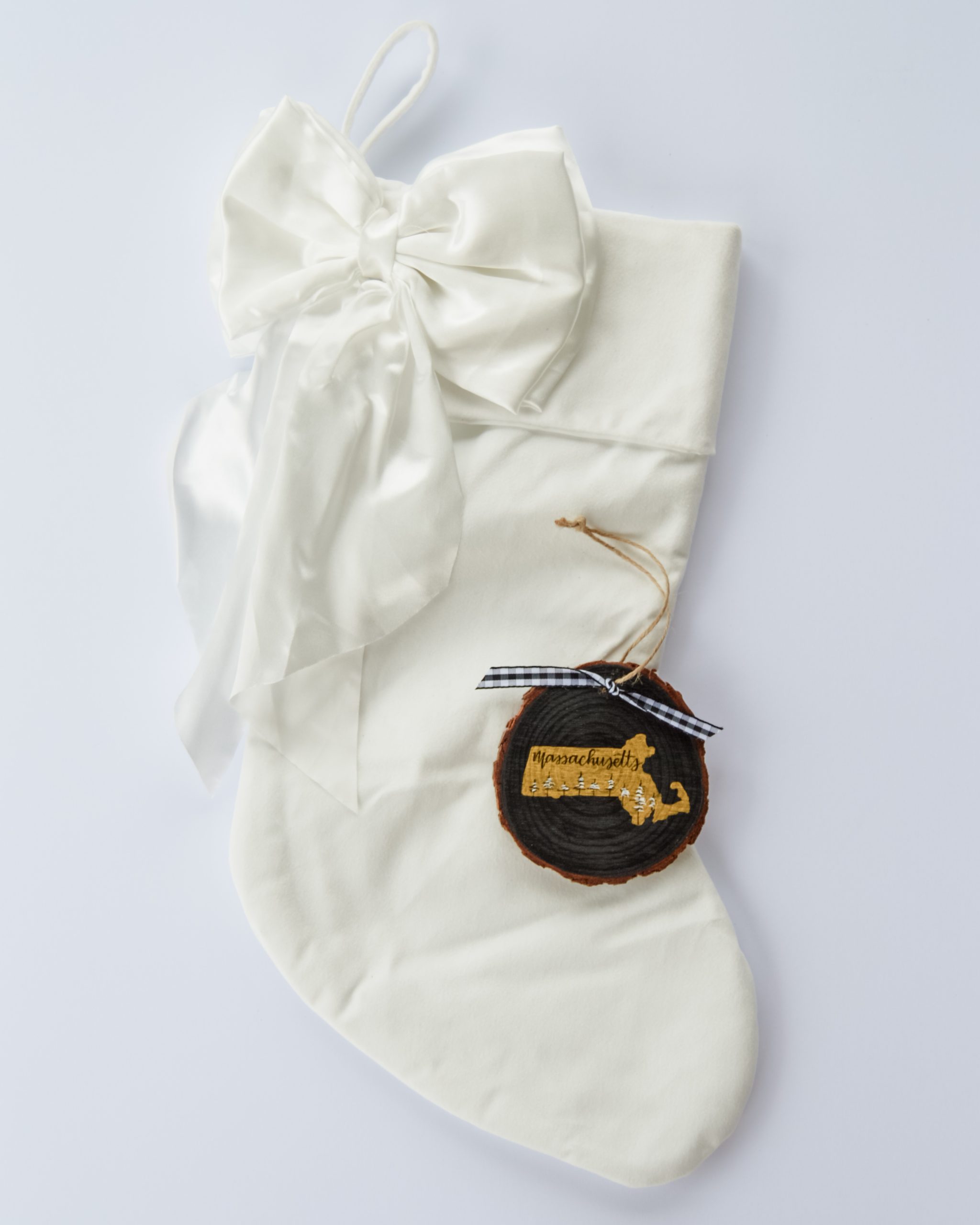 Hand-painted wood slice ornament of Massachusetts with a gold state silhouette, snow-covered trees, and the state name hand-lettered in black, tied with white buffalo plaid ribbon on a white velvet Christmas stocking