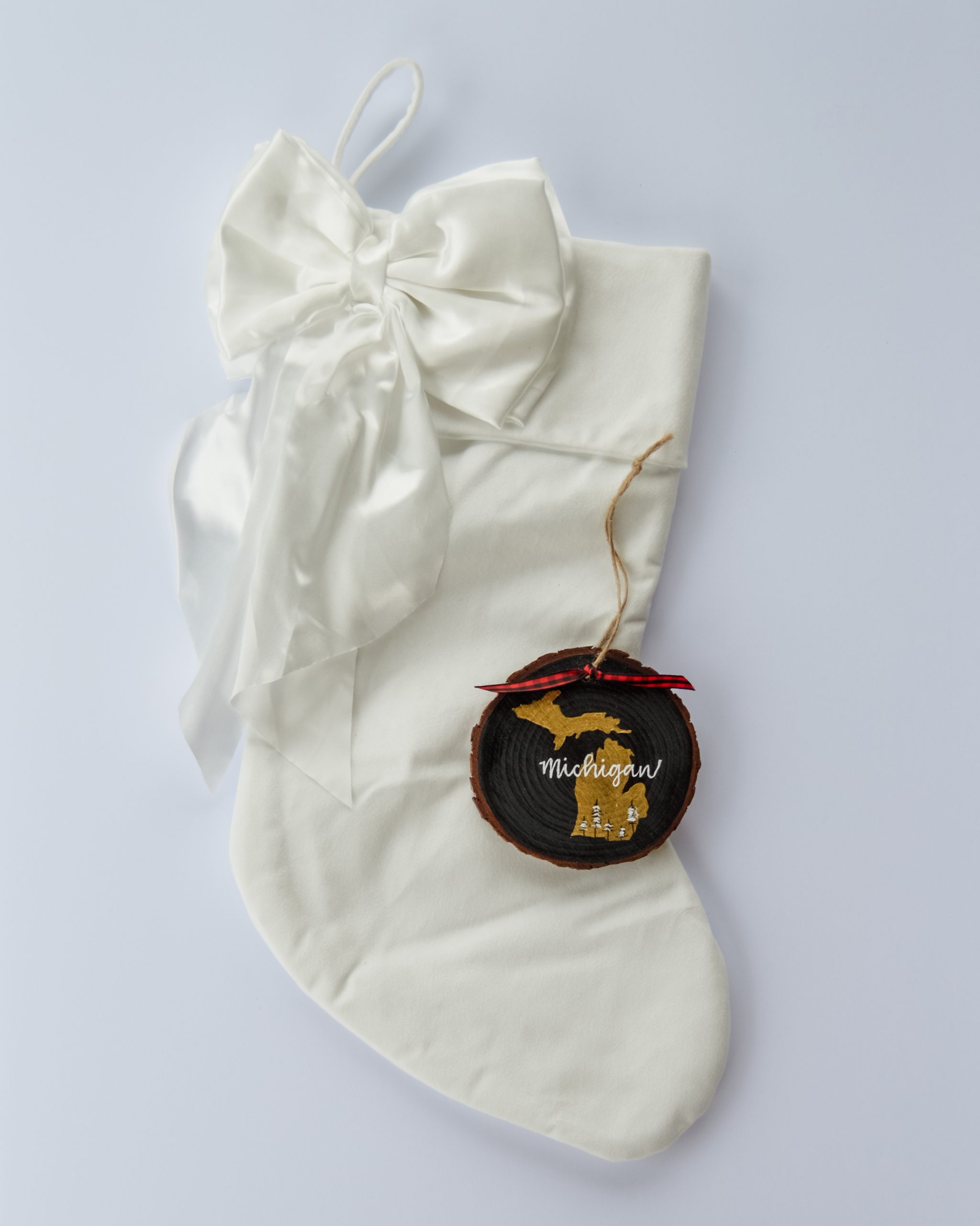 Hand-painted wood slice ornament of Michigan with a gold state silhouette, snow-covered trees, and the state name hand-lettered in white, tied with red buffalo plaid ribbon on a white velvet Christmas stocking