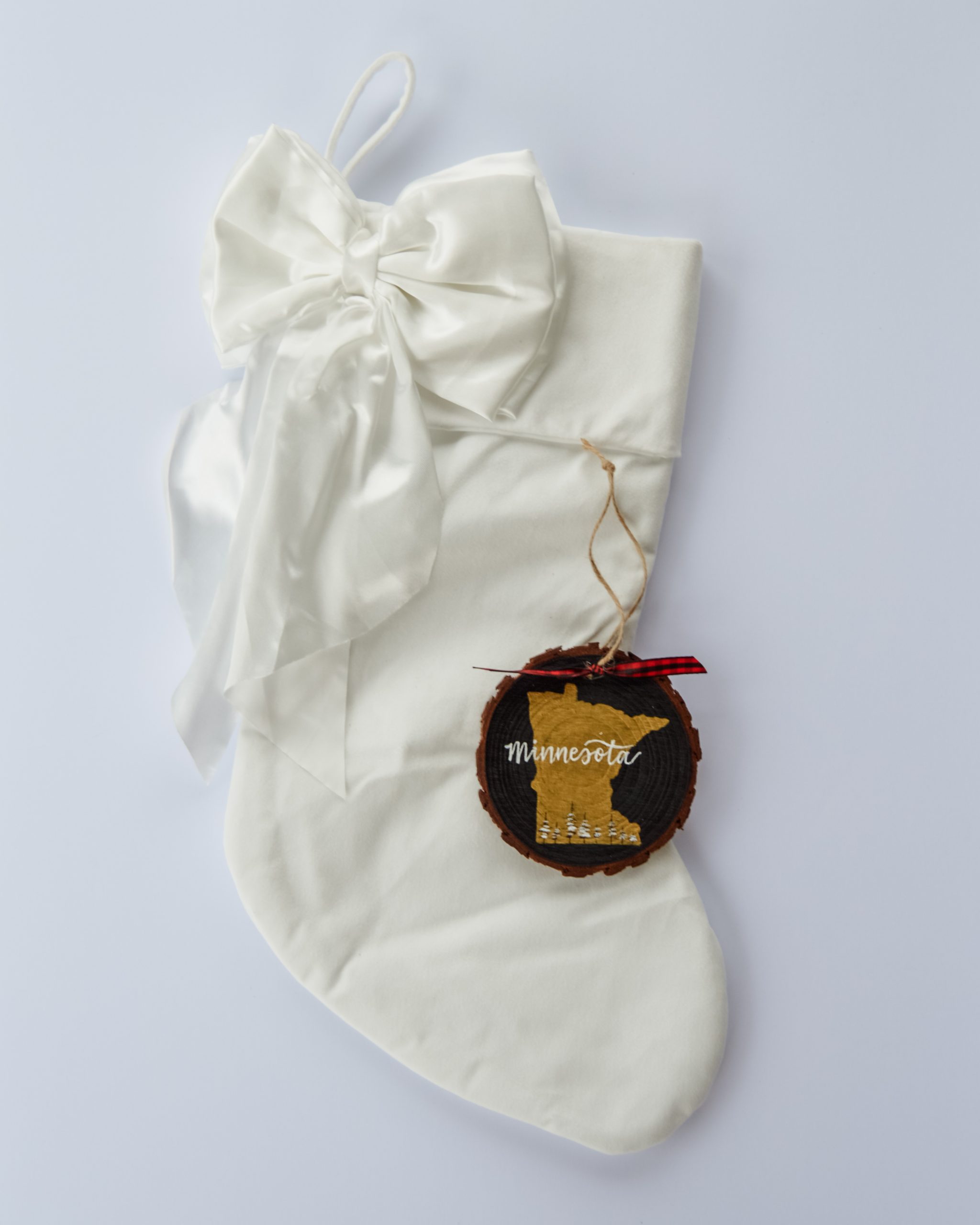 Hand-painted wood slice ornament of Minnesota with a gold state silhouette, snow-covered trees, and the state name hand-lettered in white, tied with red buffalo plaid ribbon on a white velvet Christmas stocking